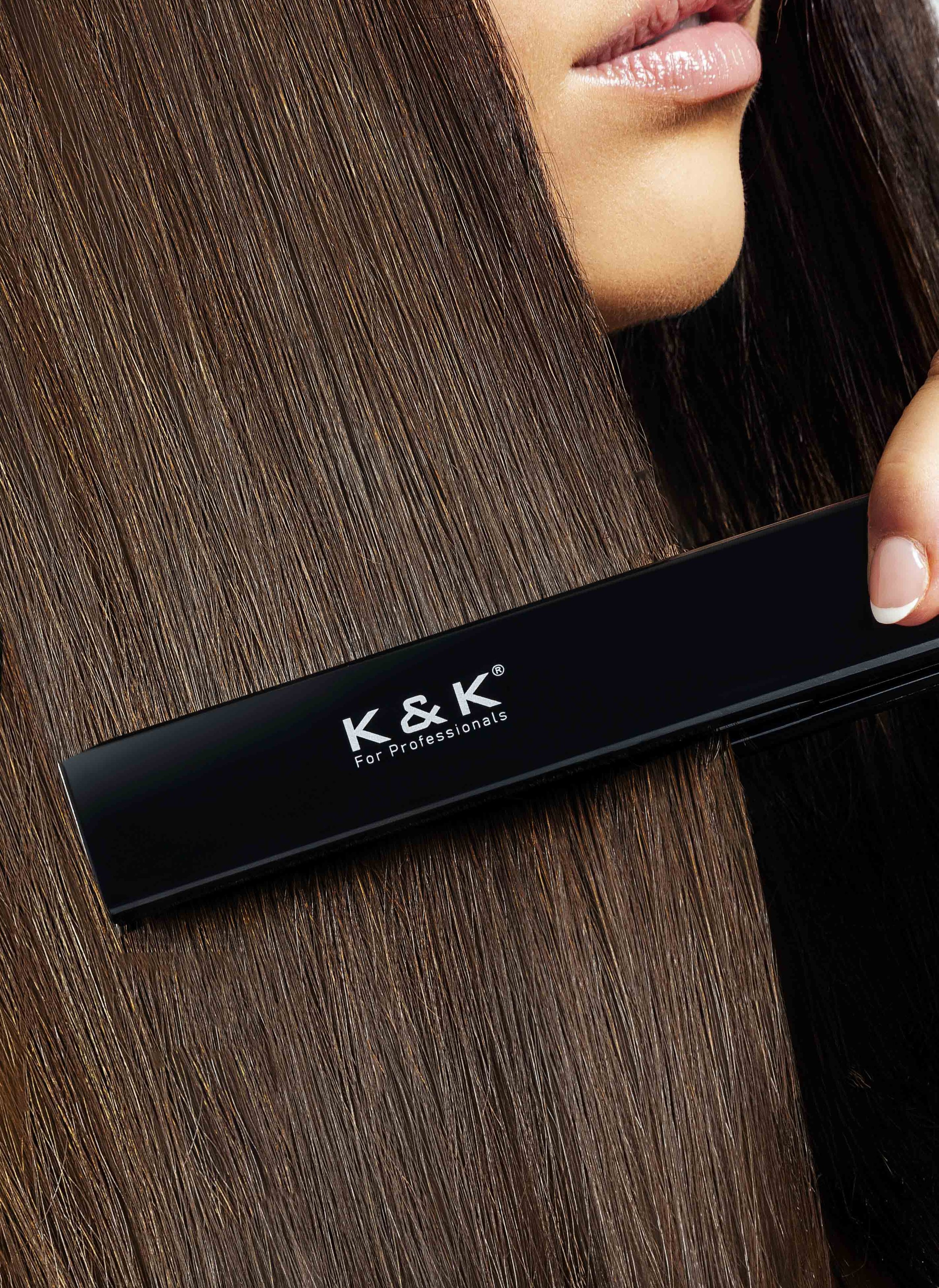 Close-up of a person using a black professional flat iron labeled 'K & K' to straighten their smooth, brown hair.