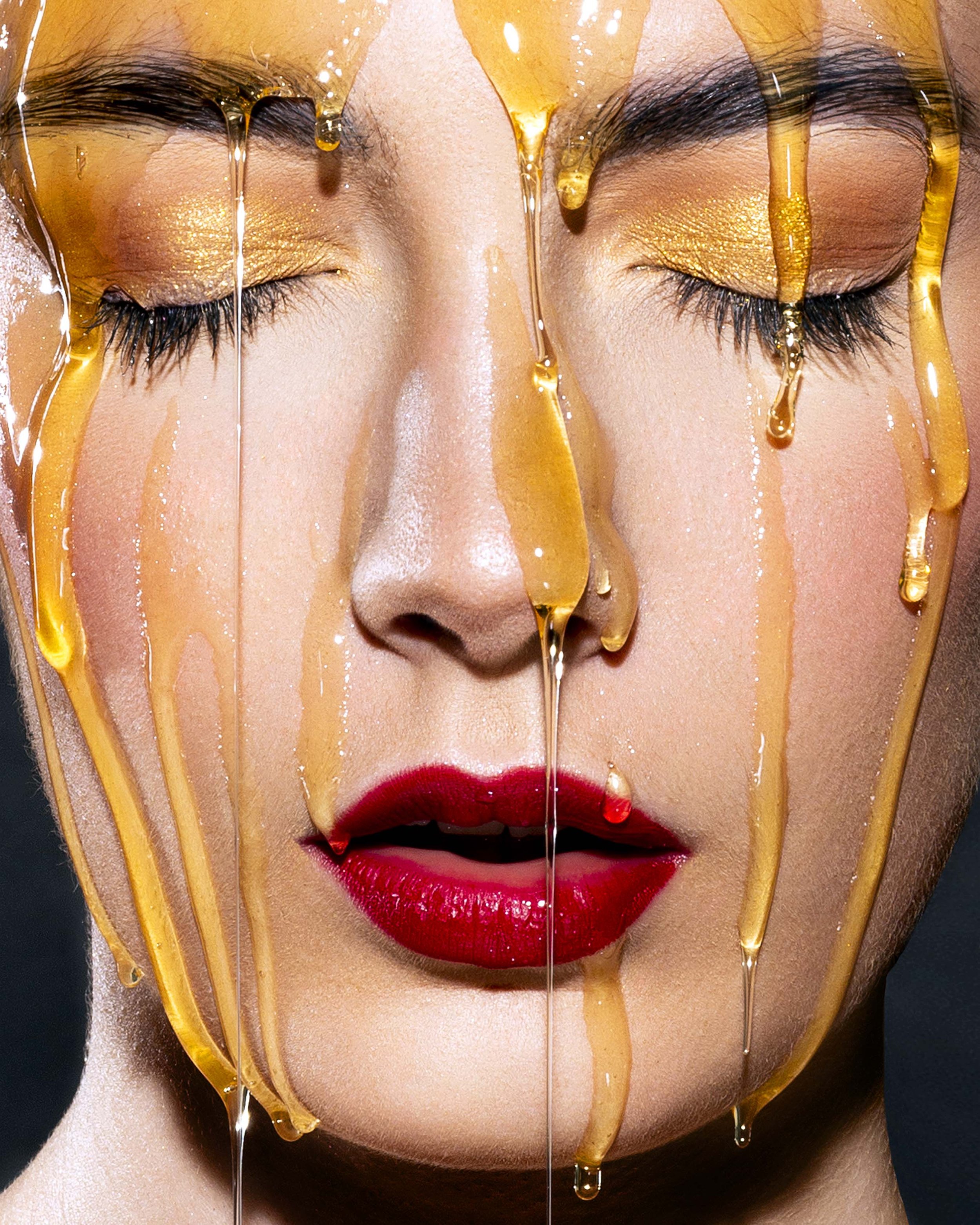 Close-up of a woman's face with red lipstick and gold eyeshadow, eyes closed, honey dripping down her face.