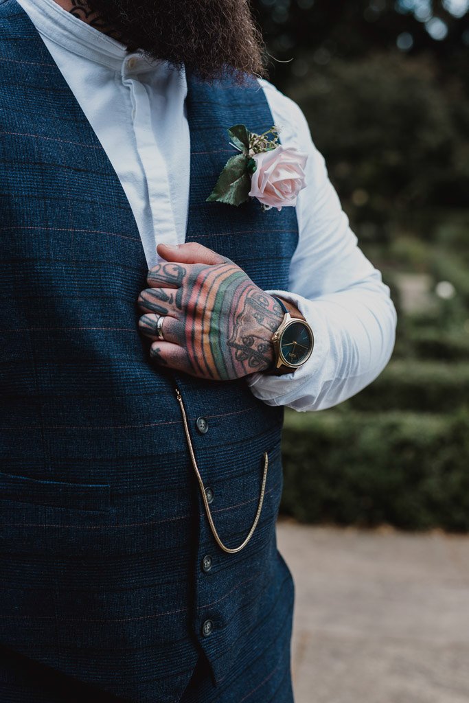 Man in a blue vest with tattoos and a watch, wearing a white shirt and a pink rose boutonniere.