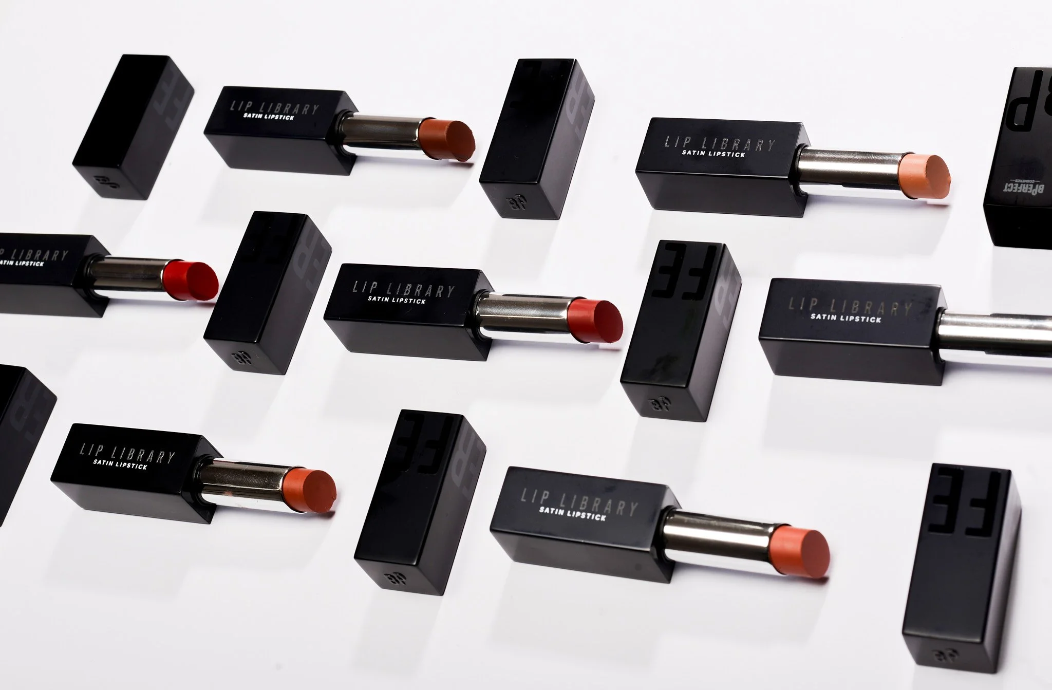 Multiple black lipstick cases labeled "Lip Library Satin Lipstick" with various lipstick shades, arranged on a white surface.