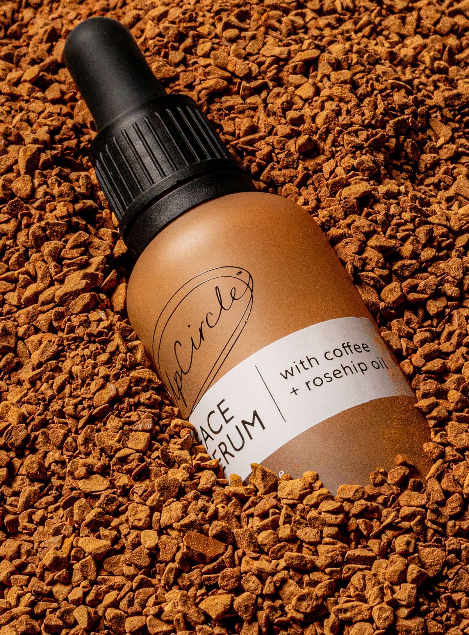 Face serum bottle with coffee and rosehip oil, surrounded by coffee grounds.