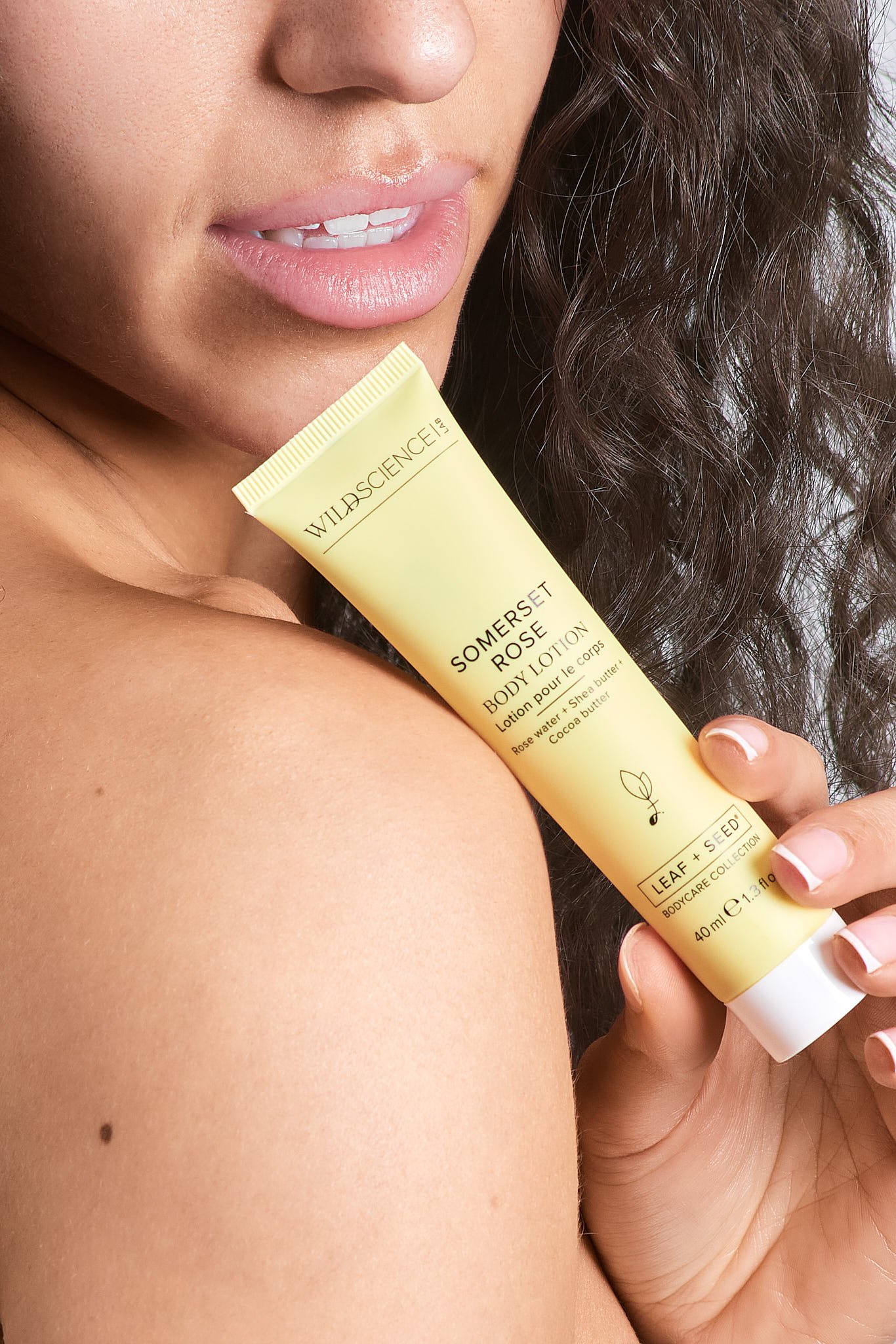 A woman with curly dark hair holding a yellow tube of Wild Science Somerset Rose Body Lotion close to her shoulder, showing her lips and part of her face.