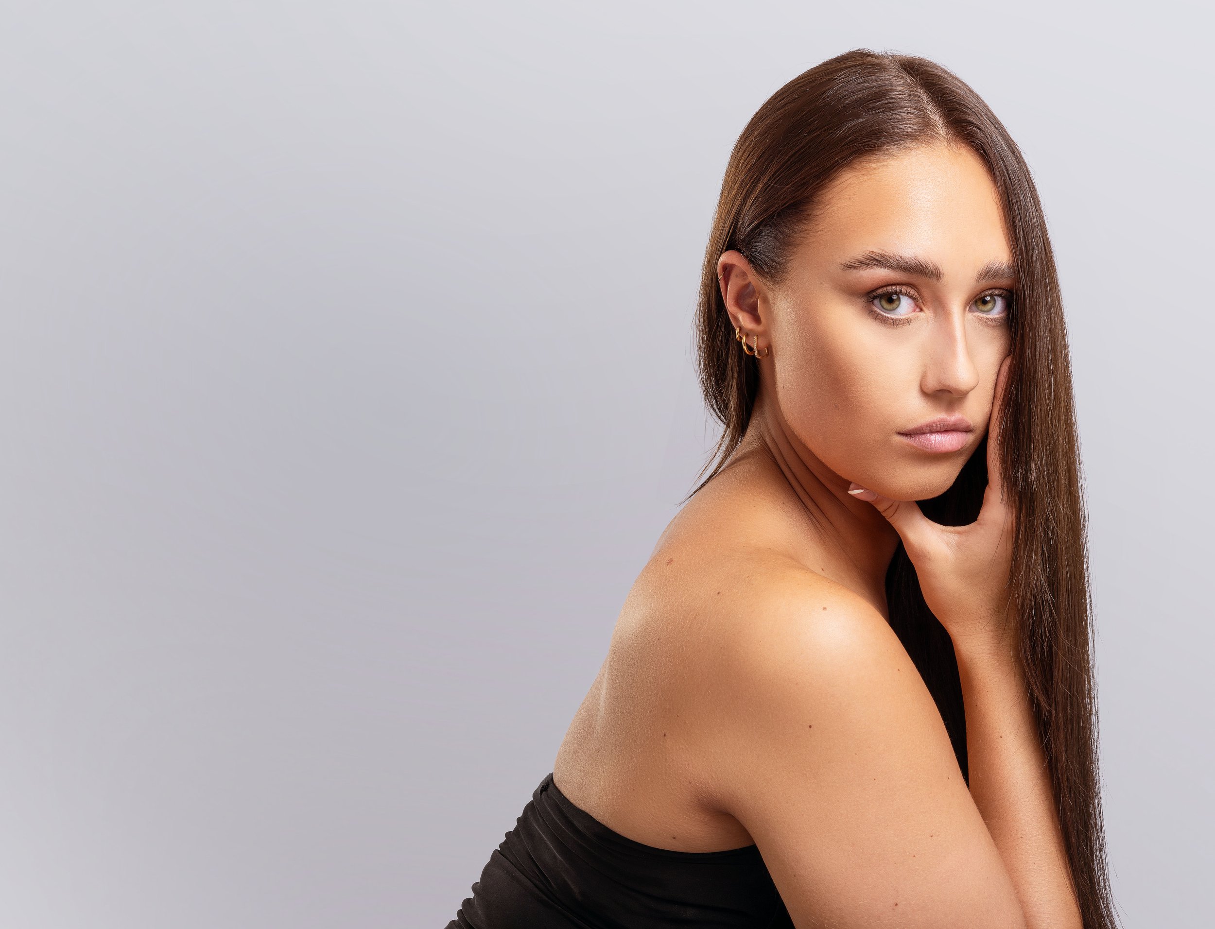 Woman with long straight brown hair, wearing a black off-shoulder top, gazing at camera, hand touching her face, neutral background.