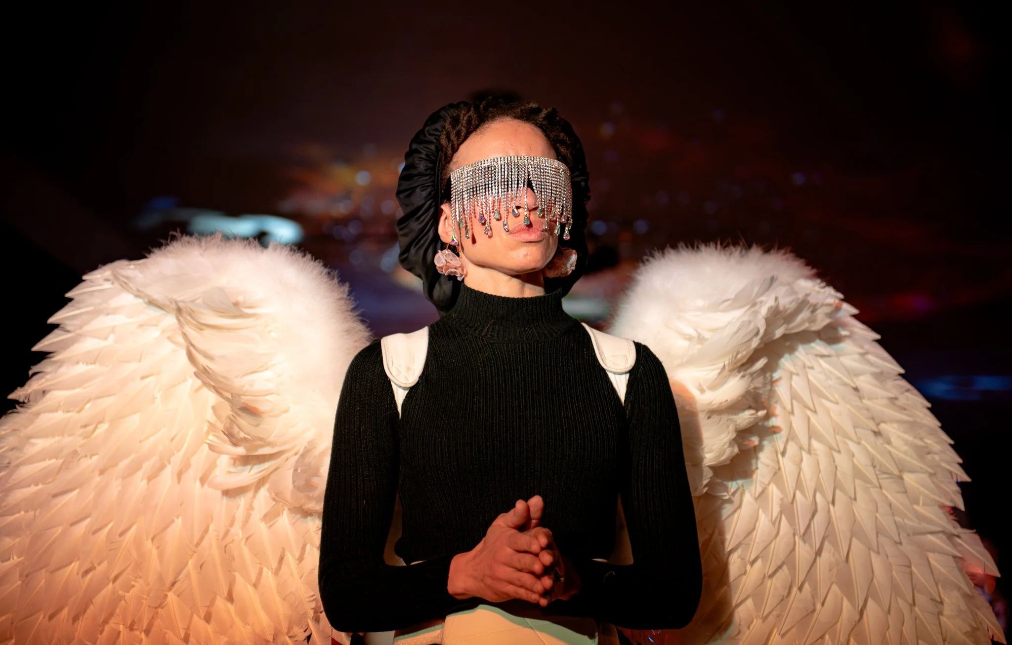 A person wearing large white feathered wings, a black turtleneck, and an elaborate jeweled face covering with hanging crystals. The individual also has earrings and a black hair bonnet.
