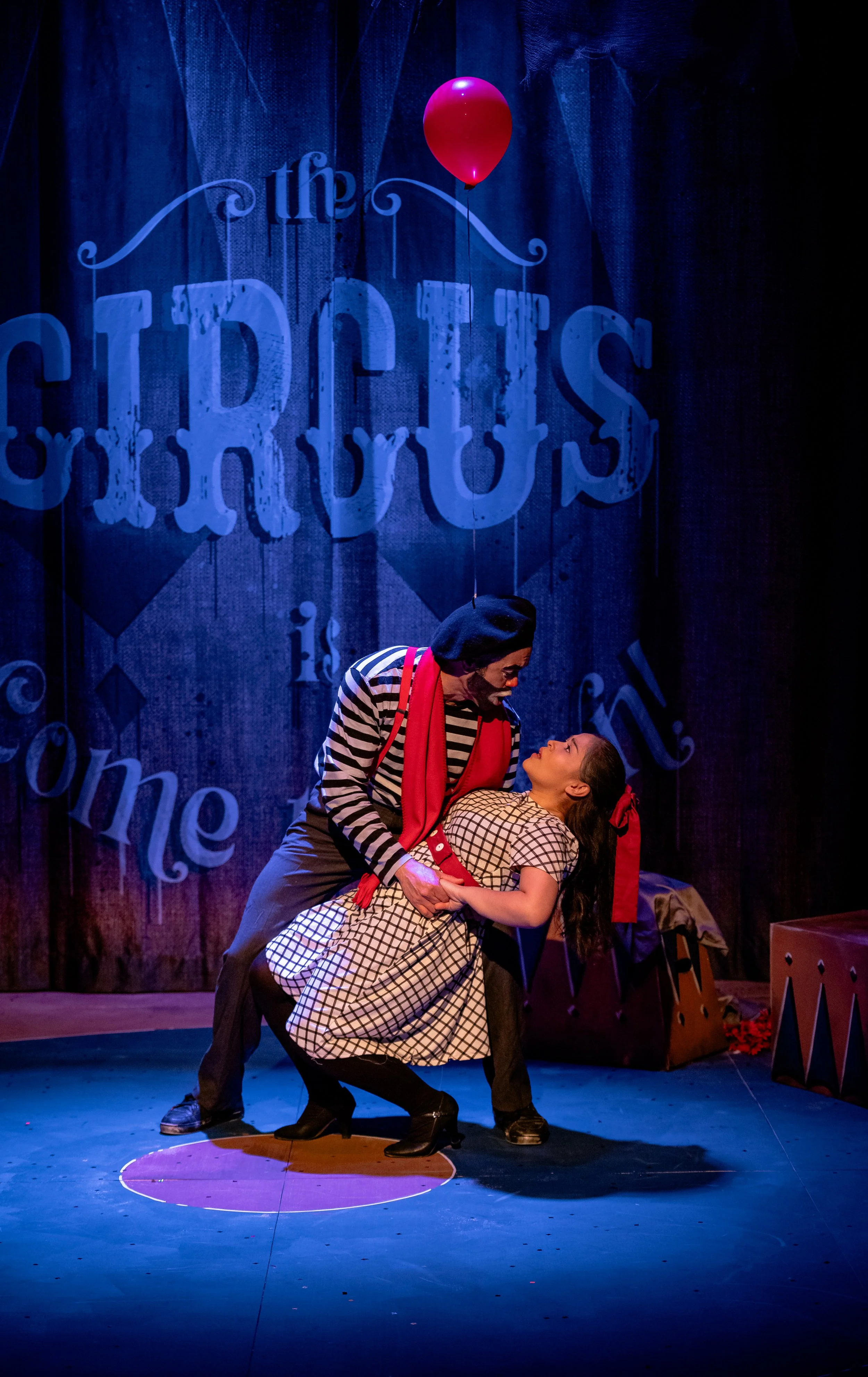 Two actors performing a scene on stage, one dressed as a clown with a beret and striped shirt, the other as a woman in a checkered dress, in front of a backdrop with the word 'the' and 'circus' on it, with a red balloon hanging above.