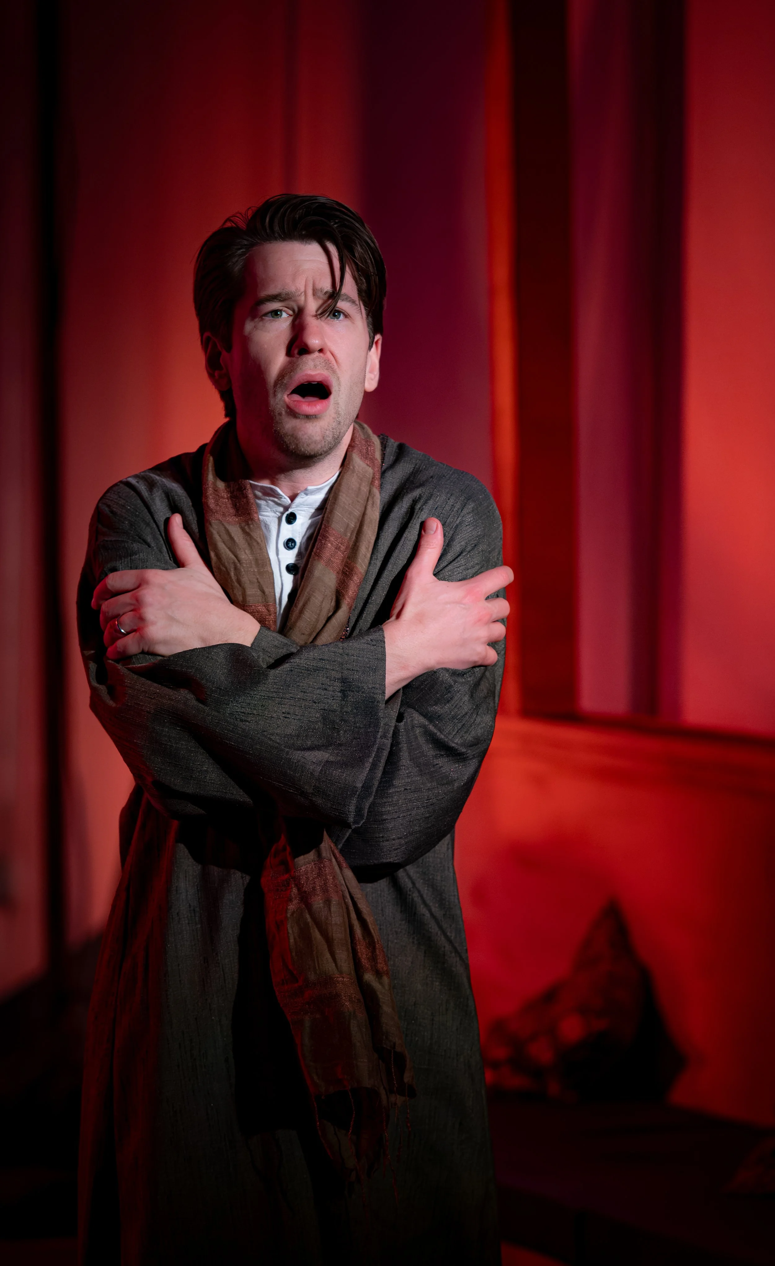 A man with dark hair, wearing a dark suit and a multicolored scarf, is standing with his arms crossed over his chest and has a distressed expression on his face. The background features red curtains.
