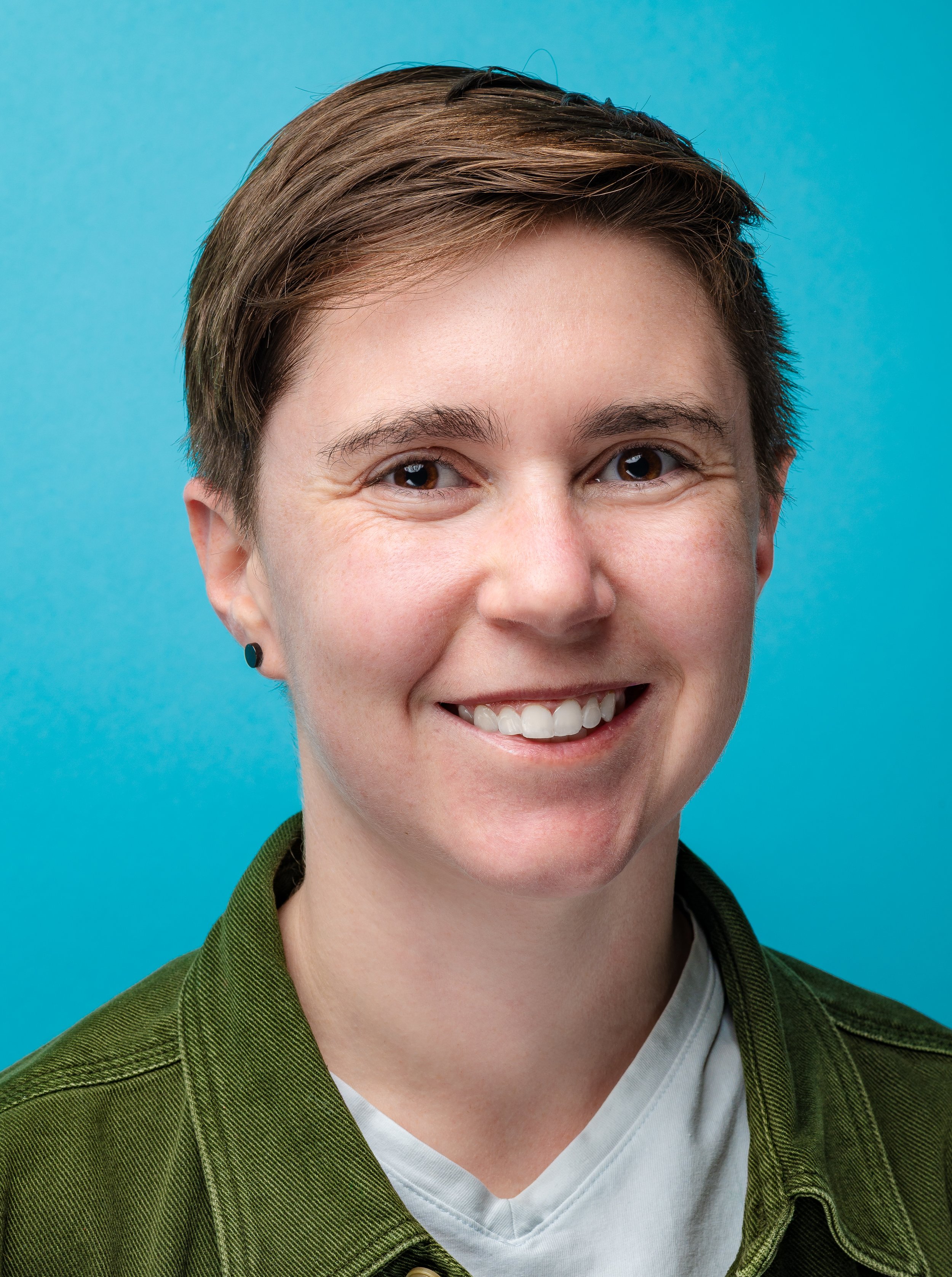Close-up of a smiling person with short brown hair, wearing a green jacket and a white shirt, against a blue background.