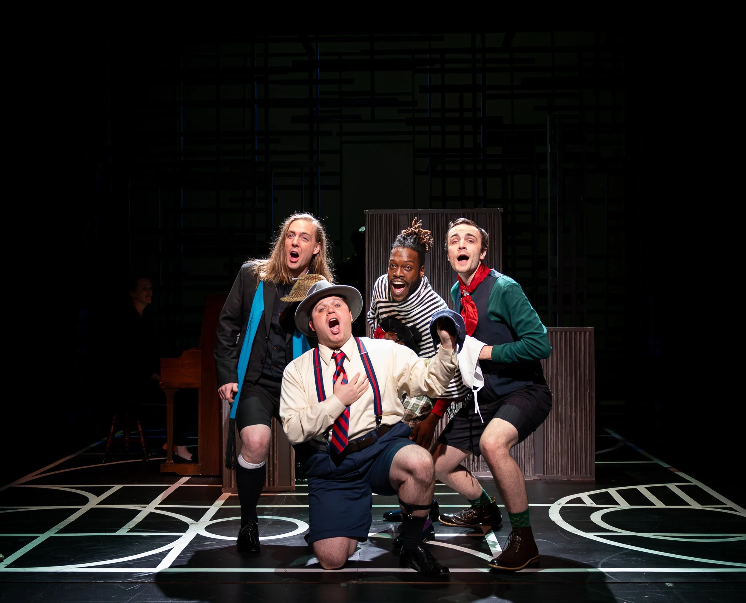 A group of five actors performing in a theatrical play on stage. They are dressed in colorful costumes and are expressive, with some singing or shouting. The background is dark with a geometric set. The stage floor has bold black and white patterns.