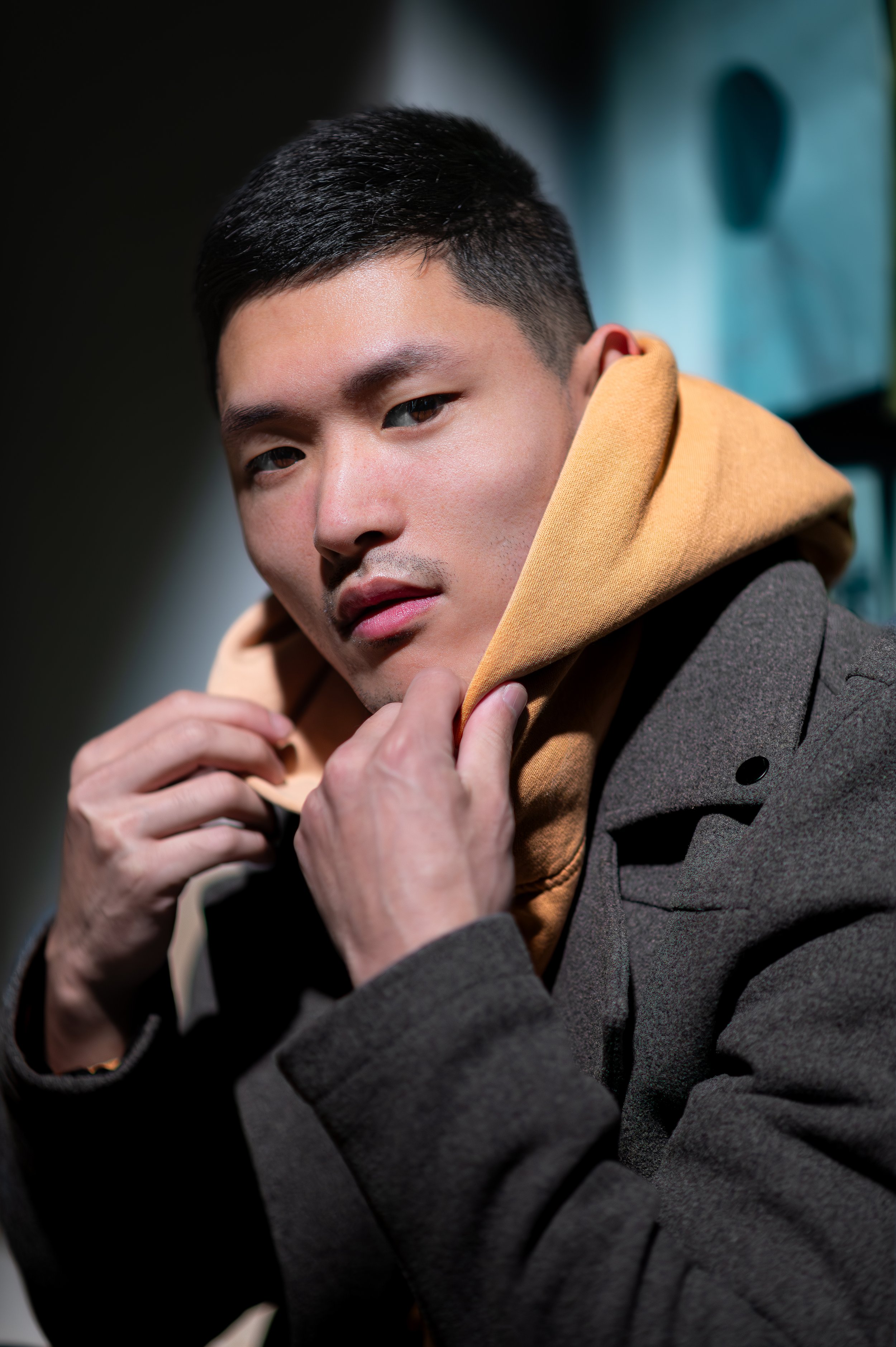 A young man with short black hair and light skin, wearing a gray coat and a tan hoodie, looking at the camera and adjusting his hoodie with his right hand indoors.