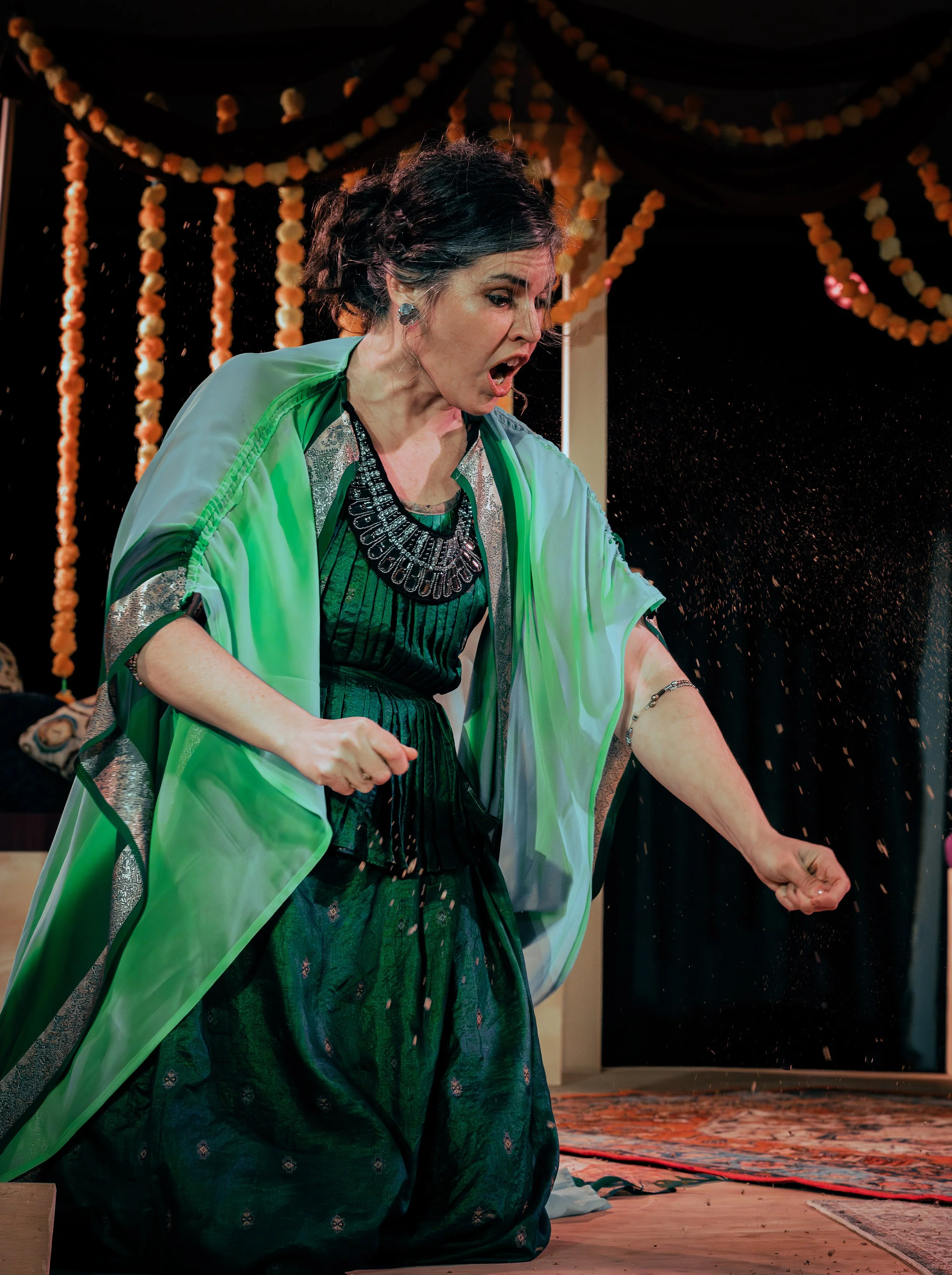 Woman in green and black traditional dress with intricate necklace, her mouth open, appearing surprised or shouting, sitting on a wooden floor with a dark background decorated with orange and white floral garlands.