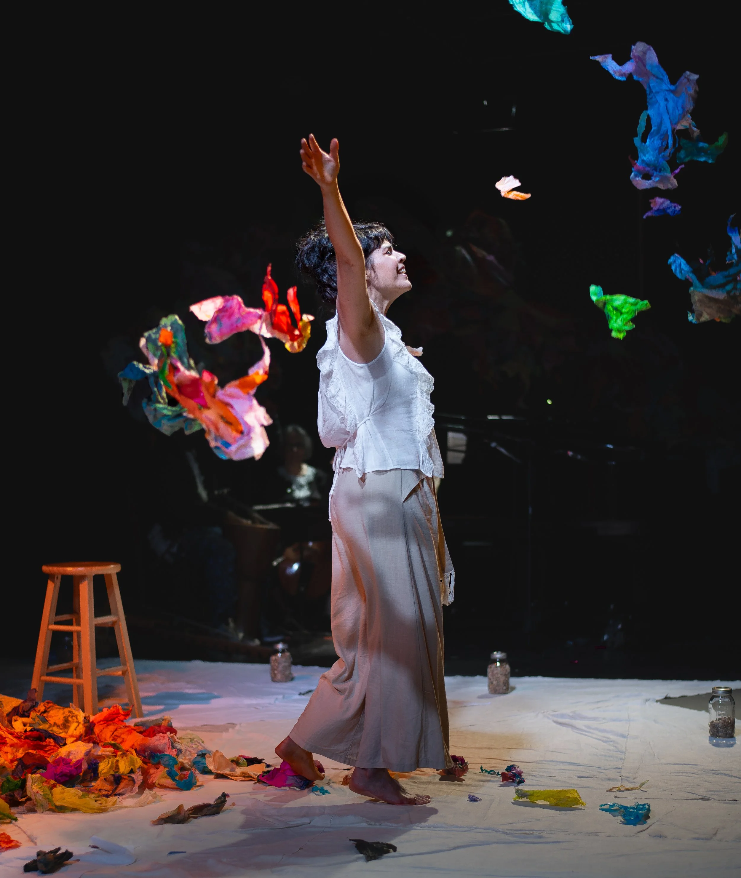 A woman with short dark hair and wearing a white sleeveless top and long beige skirt is performing on stage surrounded by colorful crumpled paper scattered on the floor. She is raising her right hand and looking upward with a joyful expression. There
