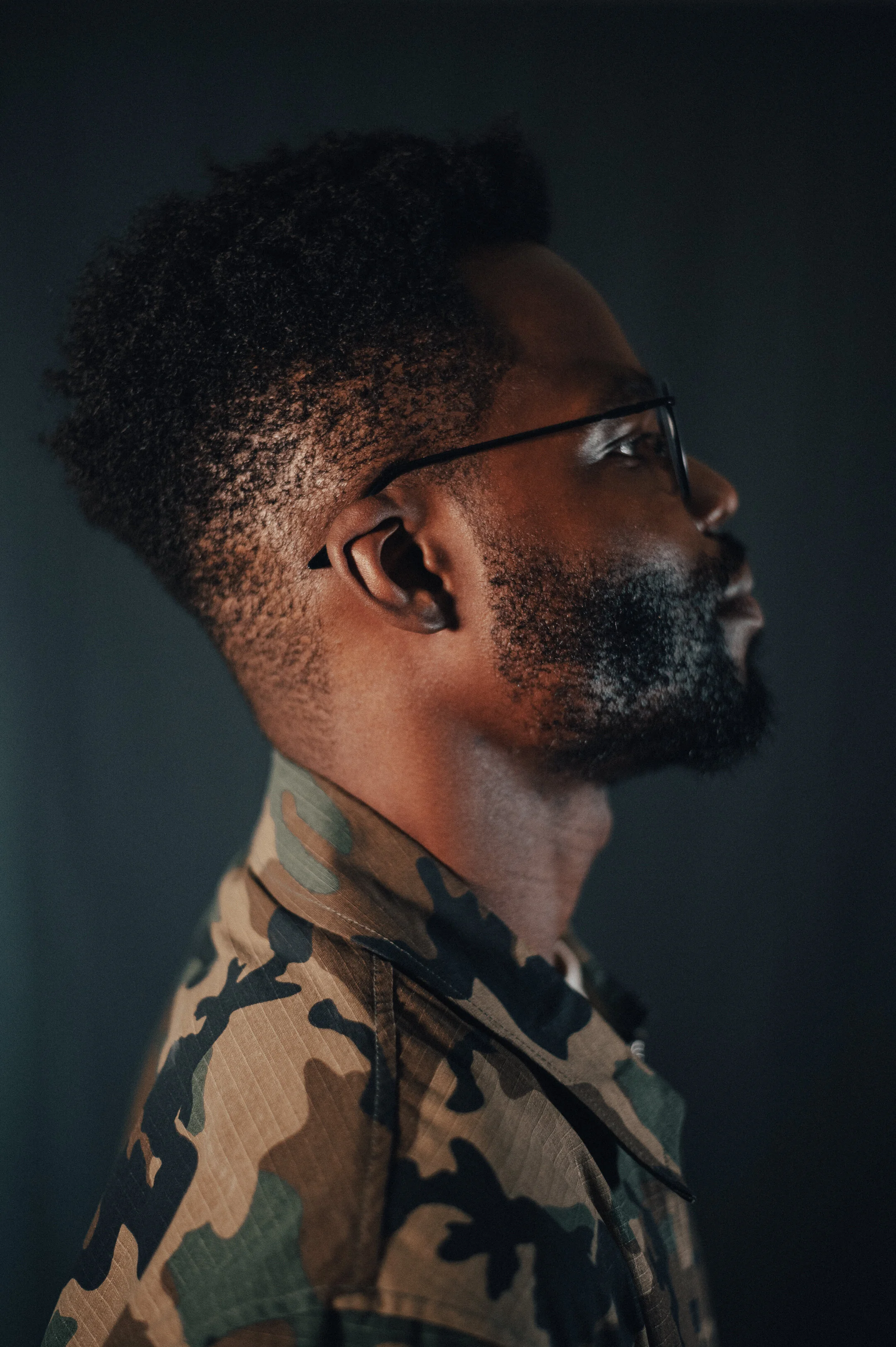 Side profile of a man with glasses, a beard, and a textured afro hairstyle, wearing a camouflage shirt against a dark background.