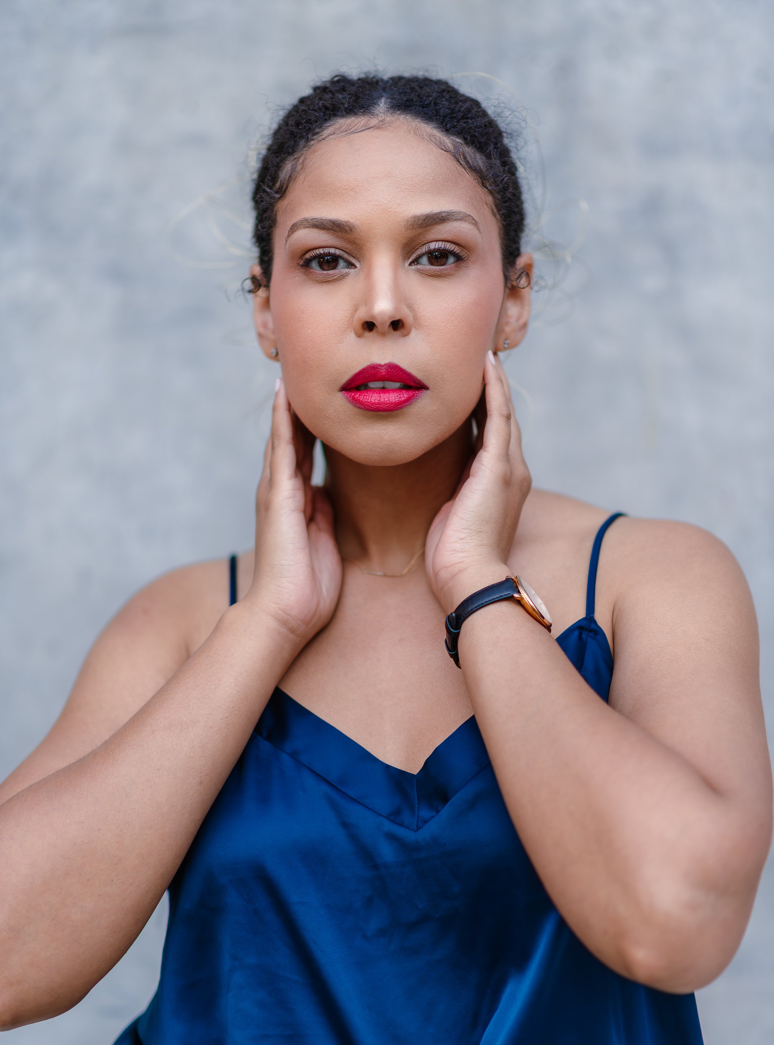 Close-up of a woman with dark curly hair, wearing red lipstick, a blue dress, and a black watch, posing with her hands near her face against a plain gray background.