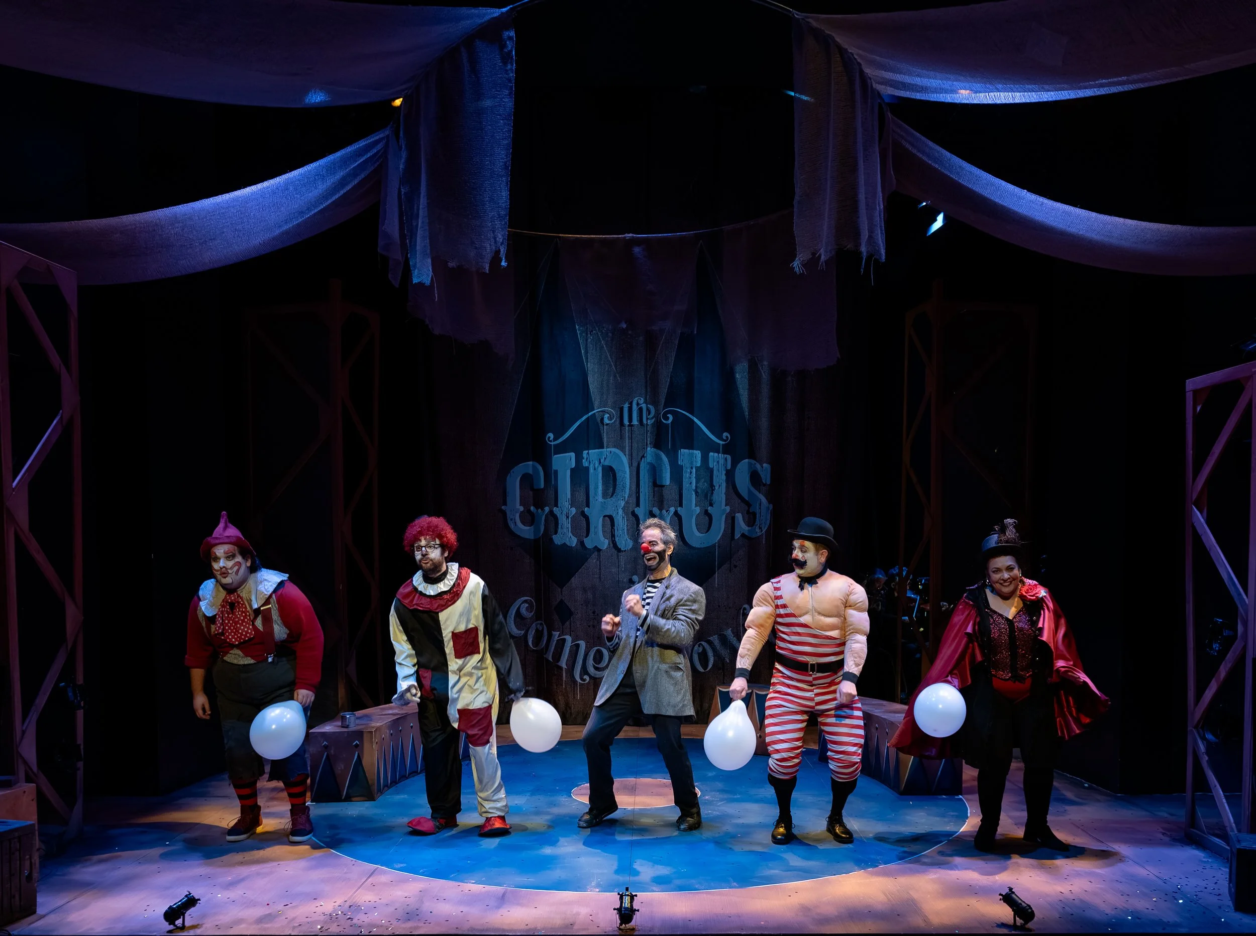 A stage show featuring five performers dressed as circus clowns and a ringmaster. The background has a sign that says 'the Circus.' The stage decor includes balloons and draped fabric overhead. The performers wear colorful costumes and makeup, and ar