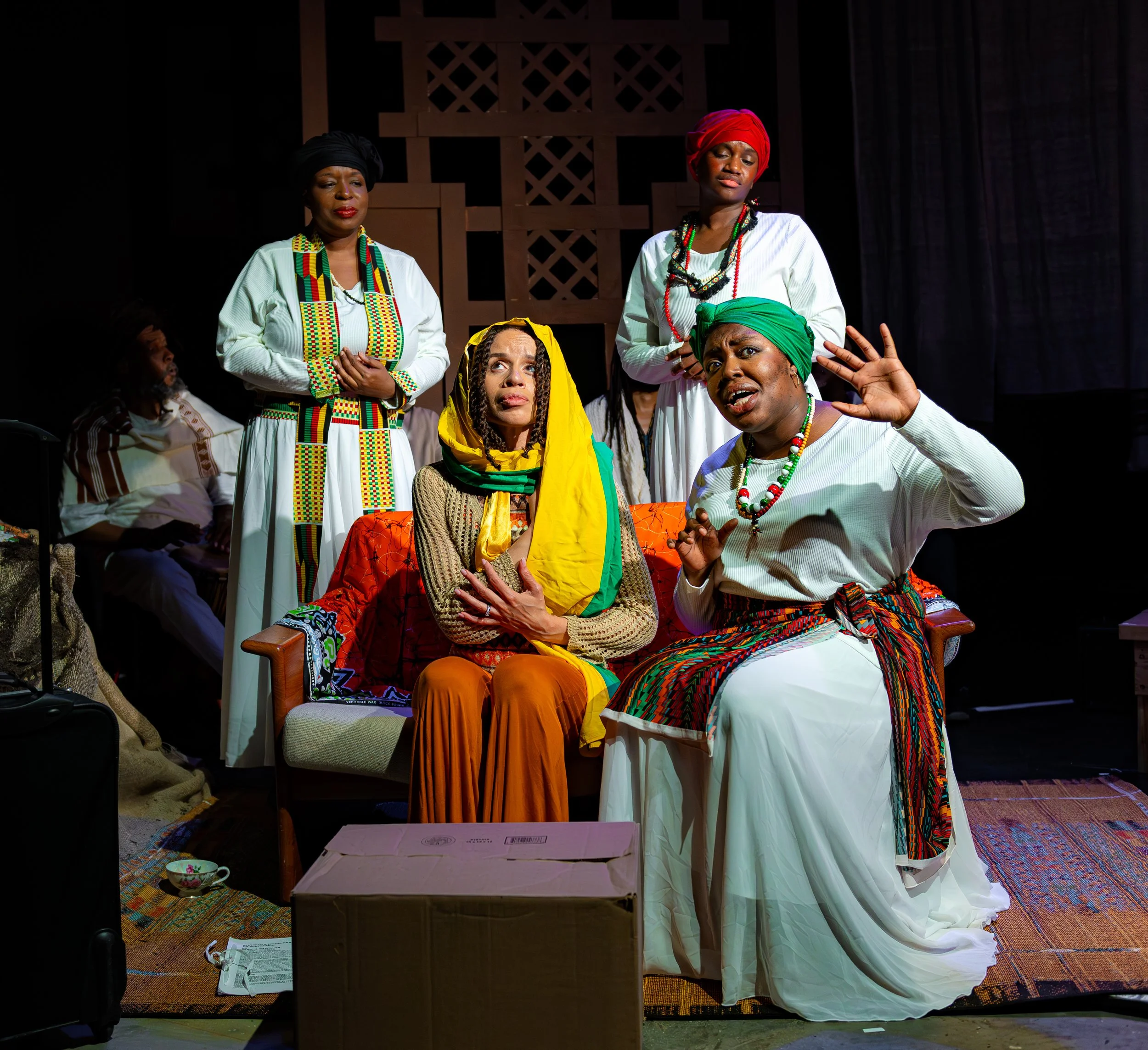 The image shows a group of African women dressed in traditional attire, participating in a cultural performance or gathering. They are wearing colorful headwraps and beaded jewelry, with some women seated and others standing. The background is dark, 