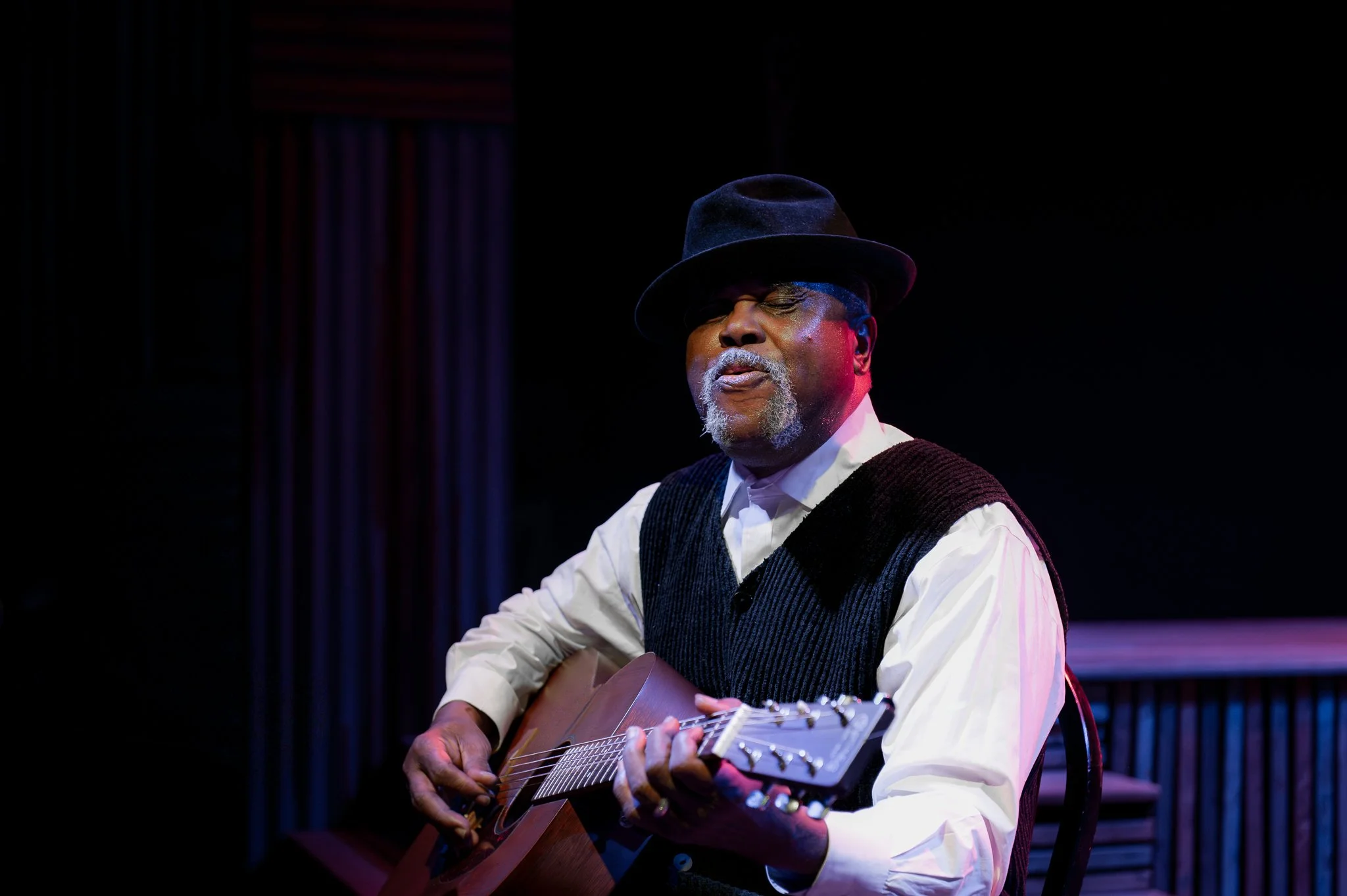 An African American man with a gray beard and mustache, wearing a black hat, playing an acoustic guitar on stage.