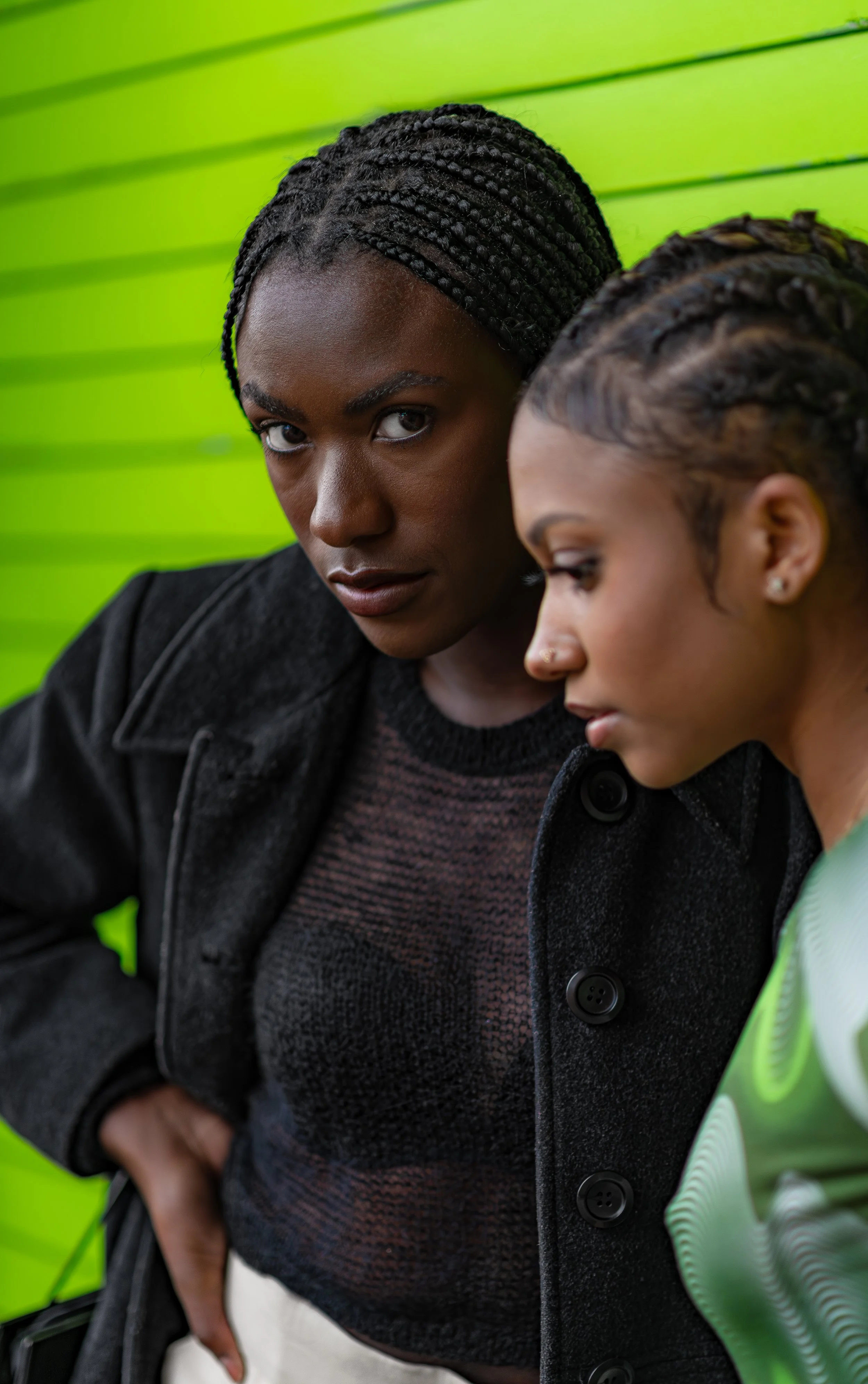 Two women with braided hair looking down with a green background.