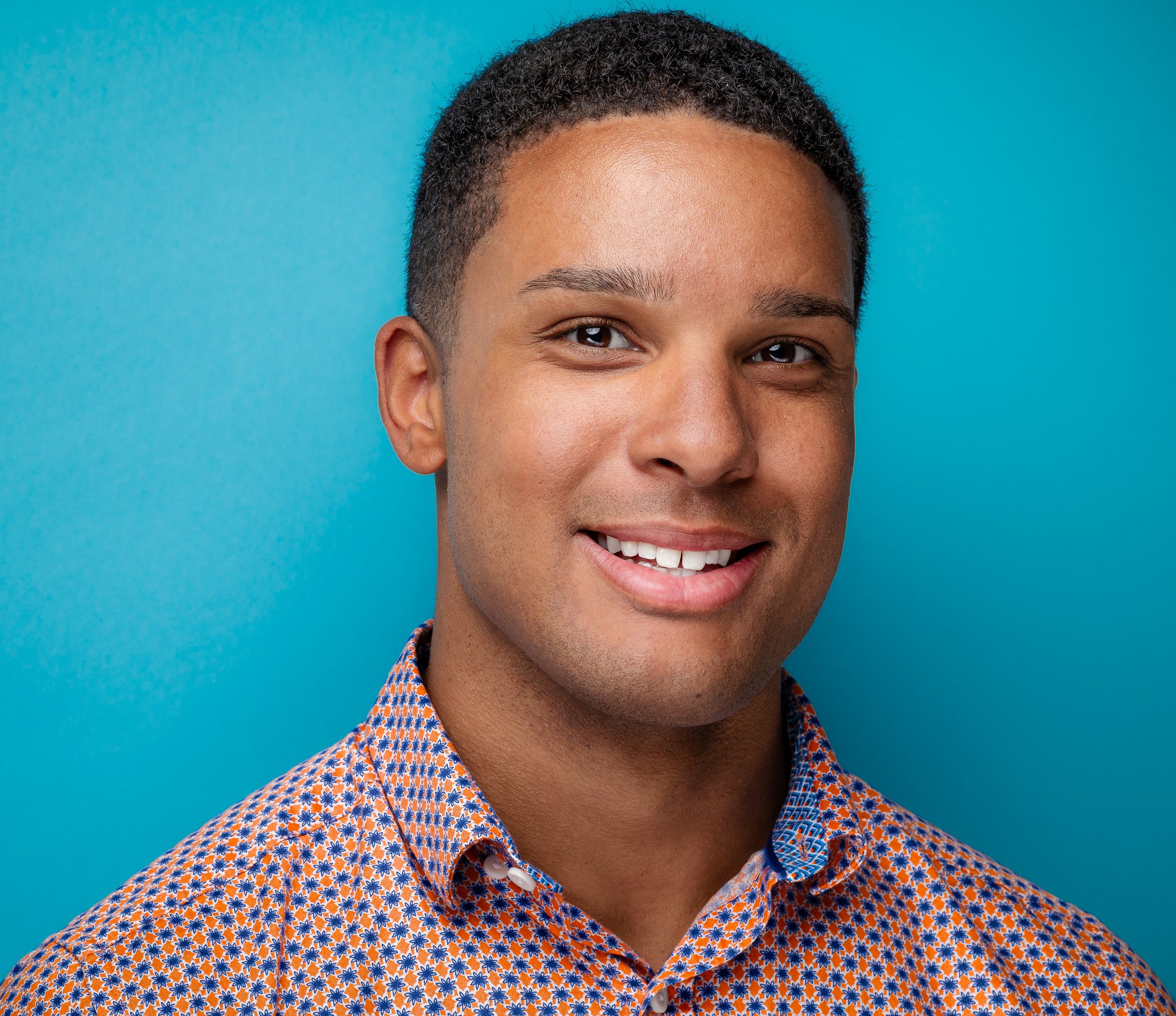 Close-up of a smiling young man with short curly hair, wearing a patterned orange and blue shirt, against a blue background.