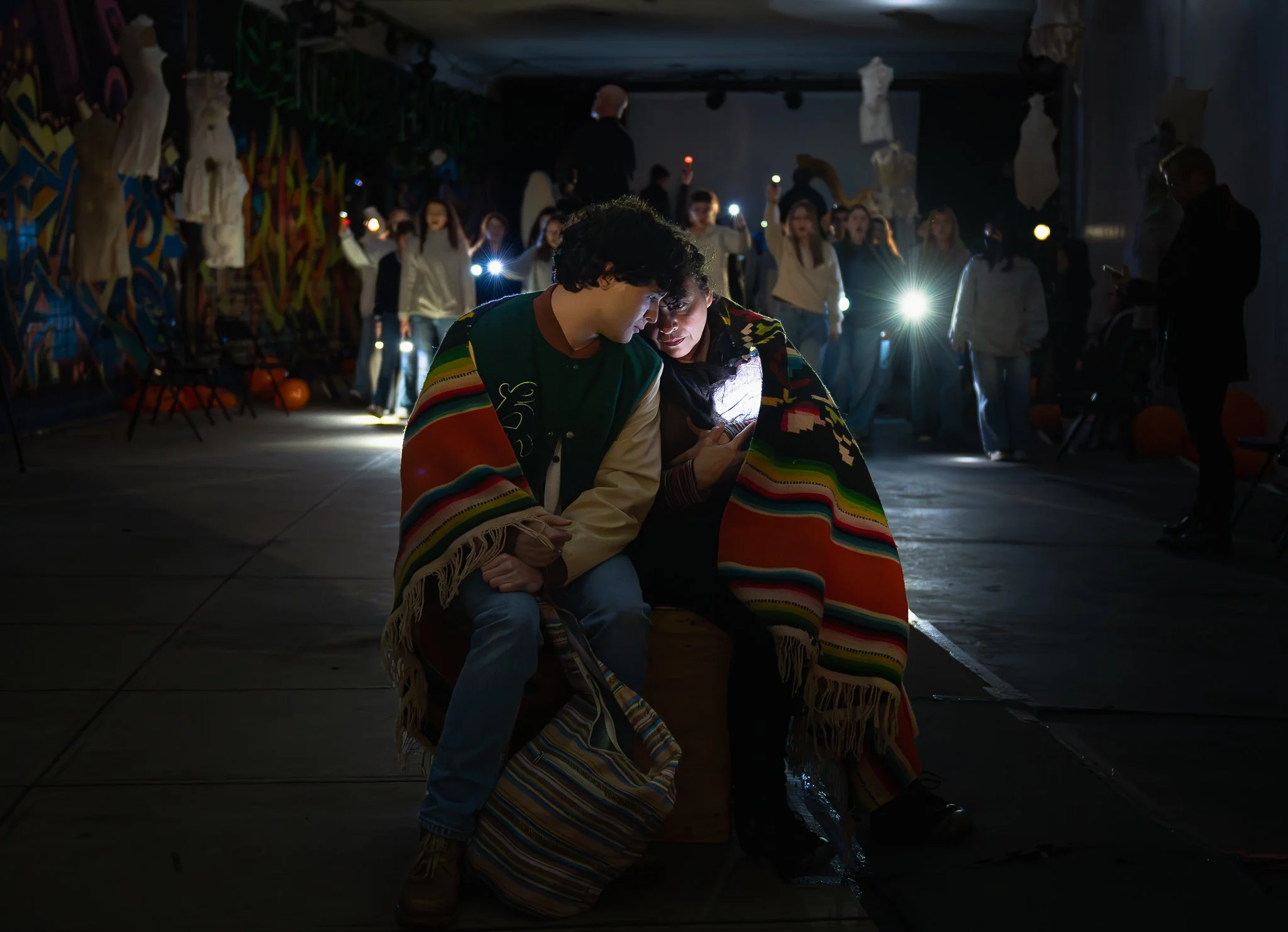 Two people crouching closely together, wearing a colorful striped blanket covering their shoulders. They are positioned on the floor with the background showing a group of people standing, holding flashlights or phone lights. The setting appears to b