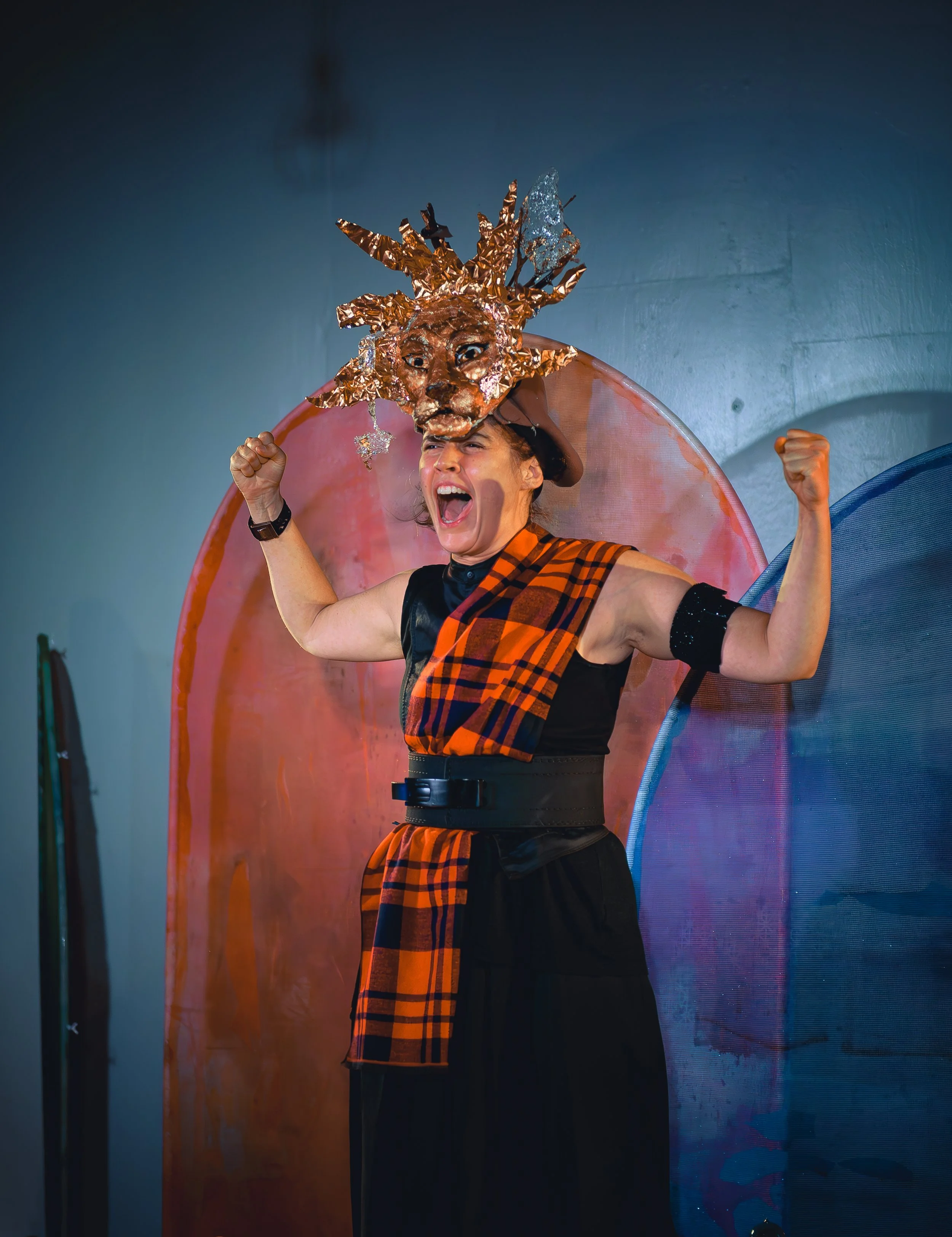 A person wearing a costume with an elaborate lion head mask made of metallic or foil material, raising their fists in excitement or victory, standing in front of colorful, translucent accordion-style backdrops.