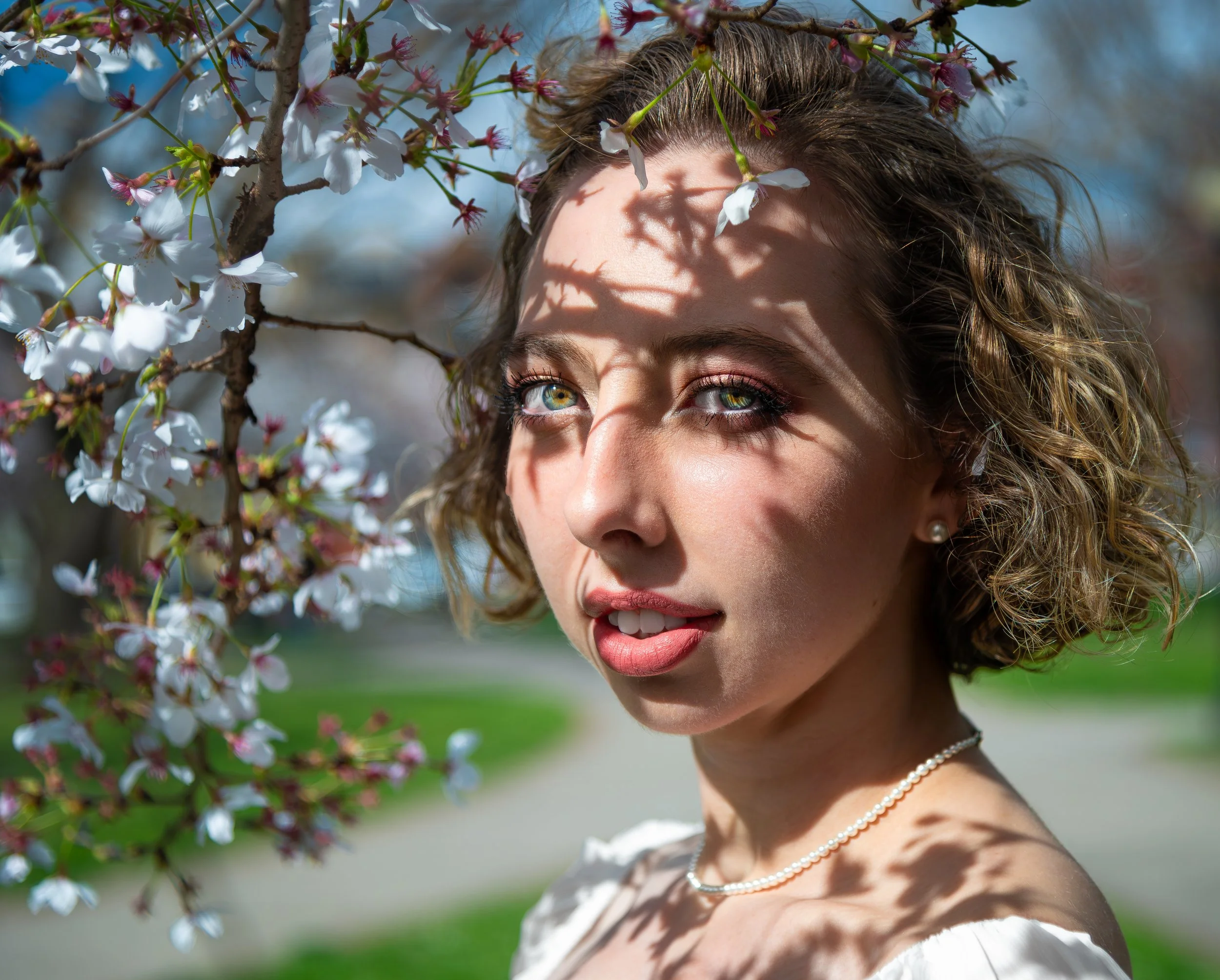 A woman with curly hair and pearl necklace smiling among blooming white cherry blossoms on a sunny day.