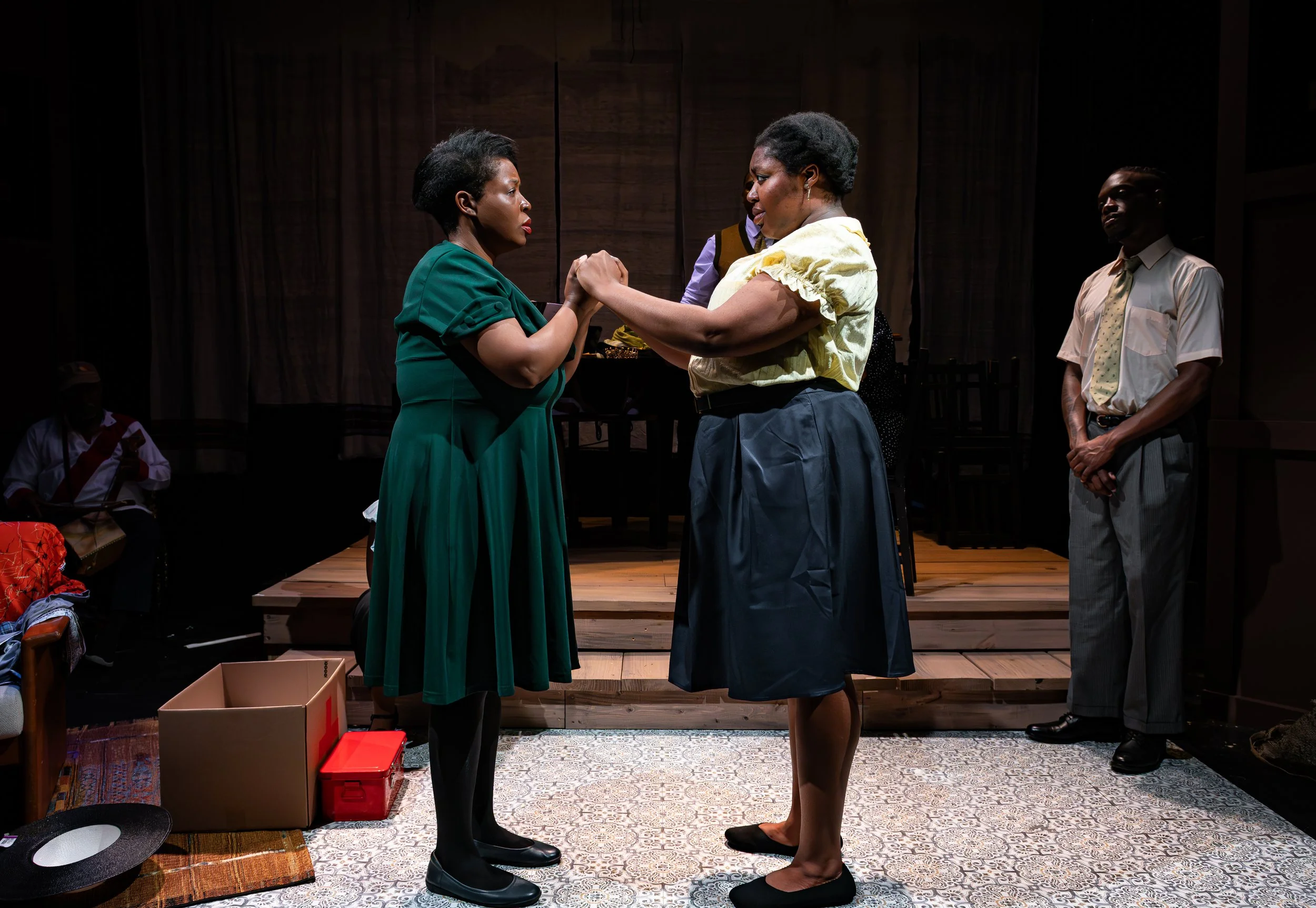 Two women holding hands and looking at each other on a stage, with three people in the background observing. One woman wears a green dress, and the other wears a light yellow blouse with a dark skirt. Stage has wood paneling and various props.