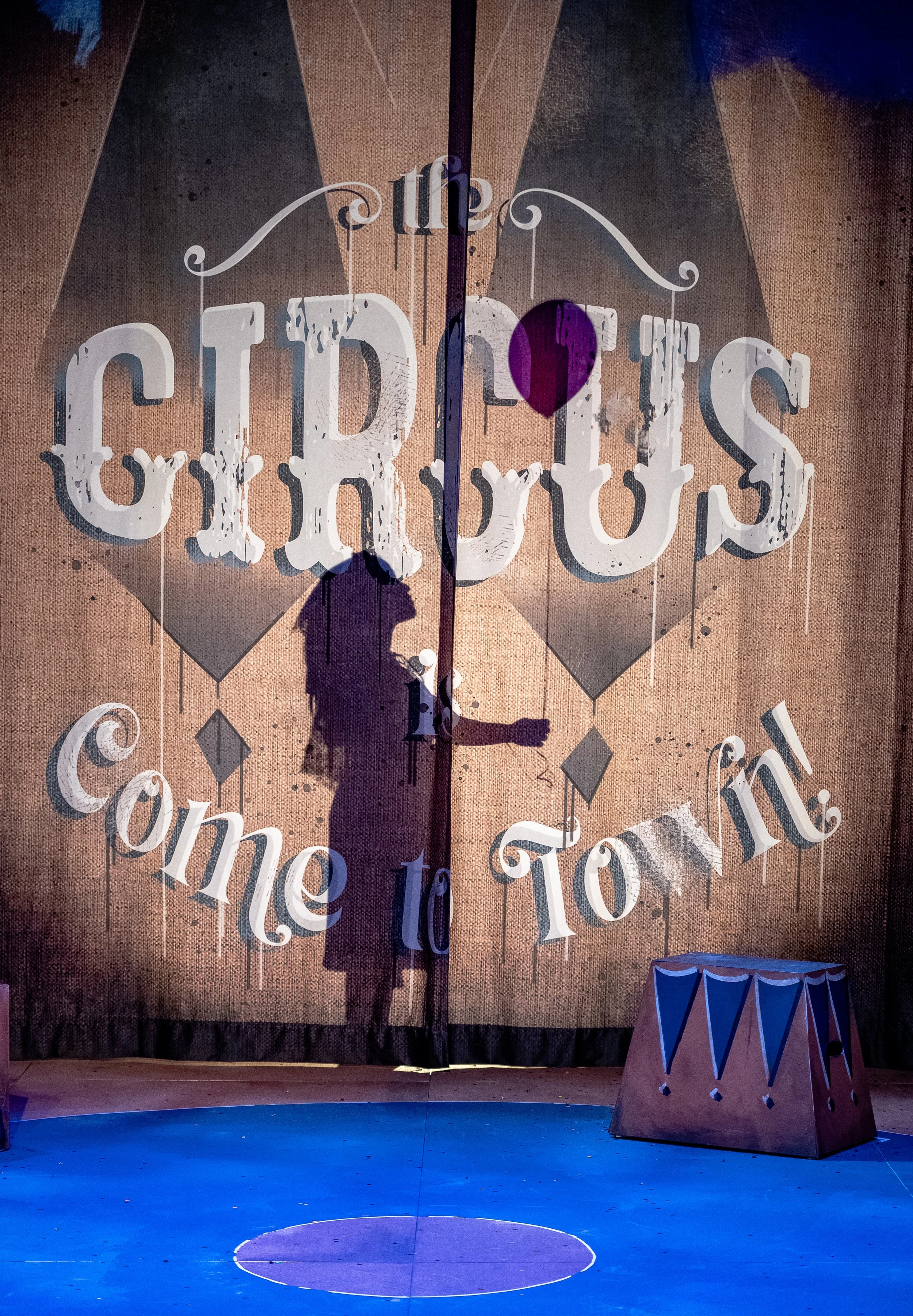 Stage with a textured backdrop displaying the words "the CIRCUS come to town" and a silhouette of a performer in a circus costume. There is a small, colorful circus-themed prop on the stage floor.