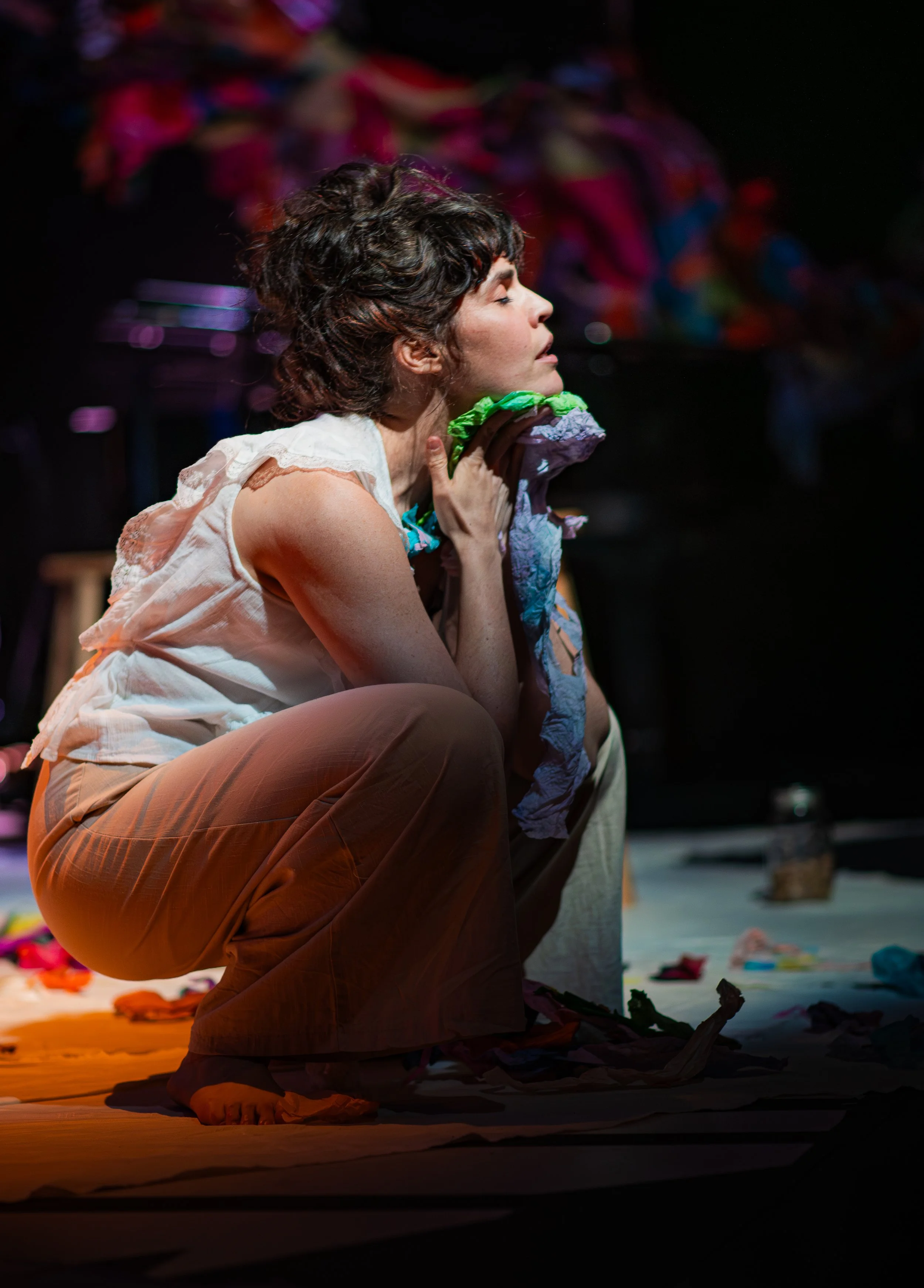 A woman with short, curly dark hair is sitting on the floor in a dark room, eyes closed, holding a piece of green fabric to her neck, with colorful fabrics in the background.