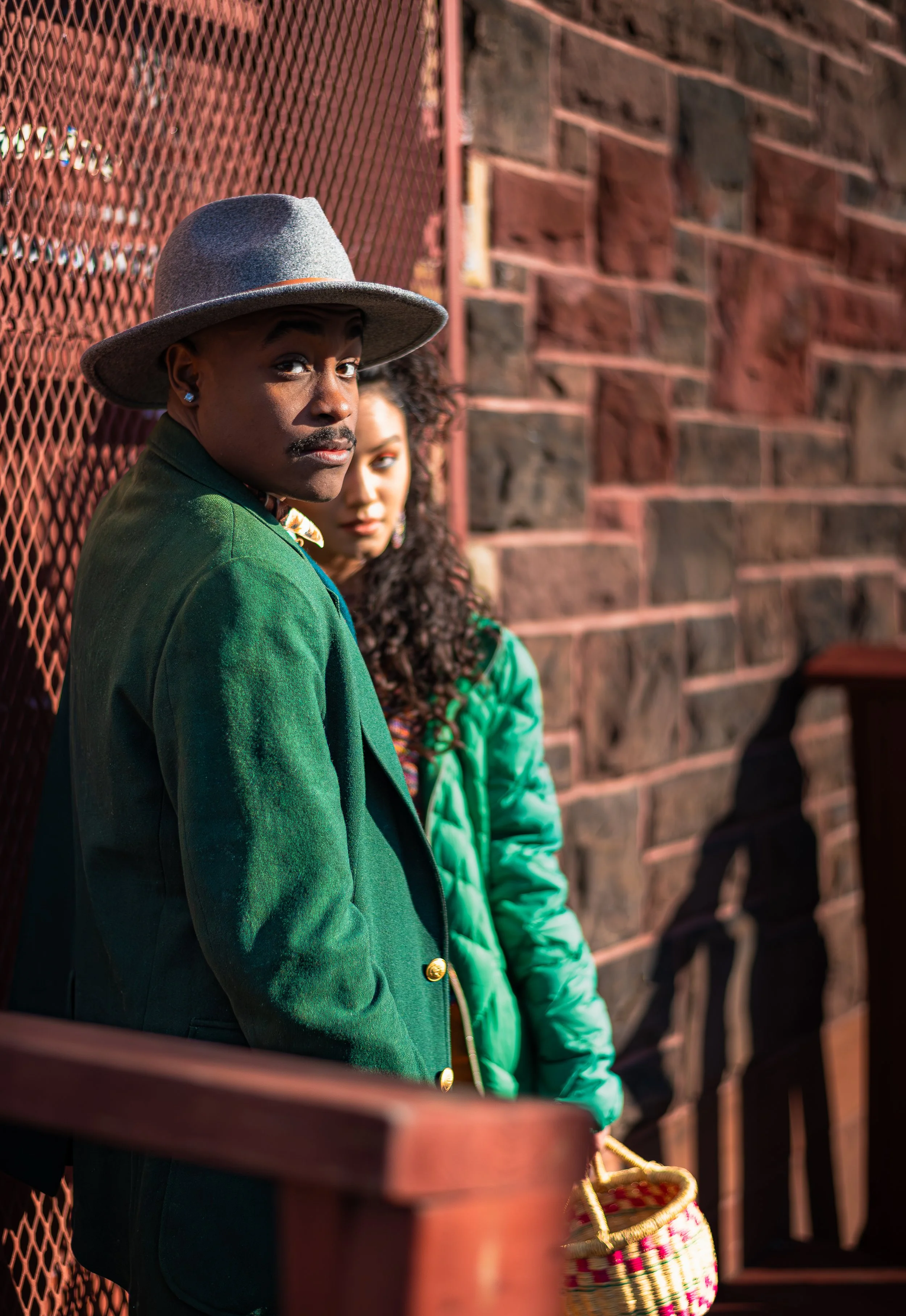 A woman in a green jacket and gray hat, standing next to a woman in a turquoise coat, against a brick wall, partially visible behind a wooden railing.