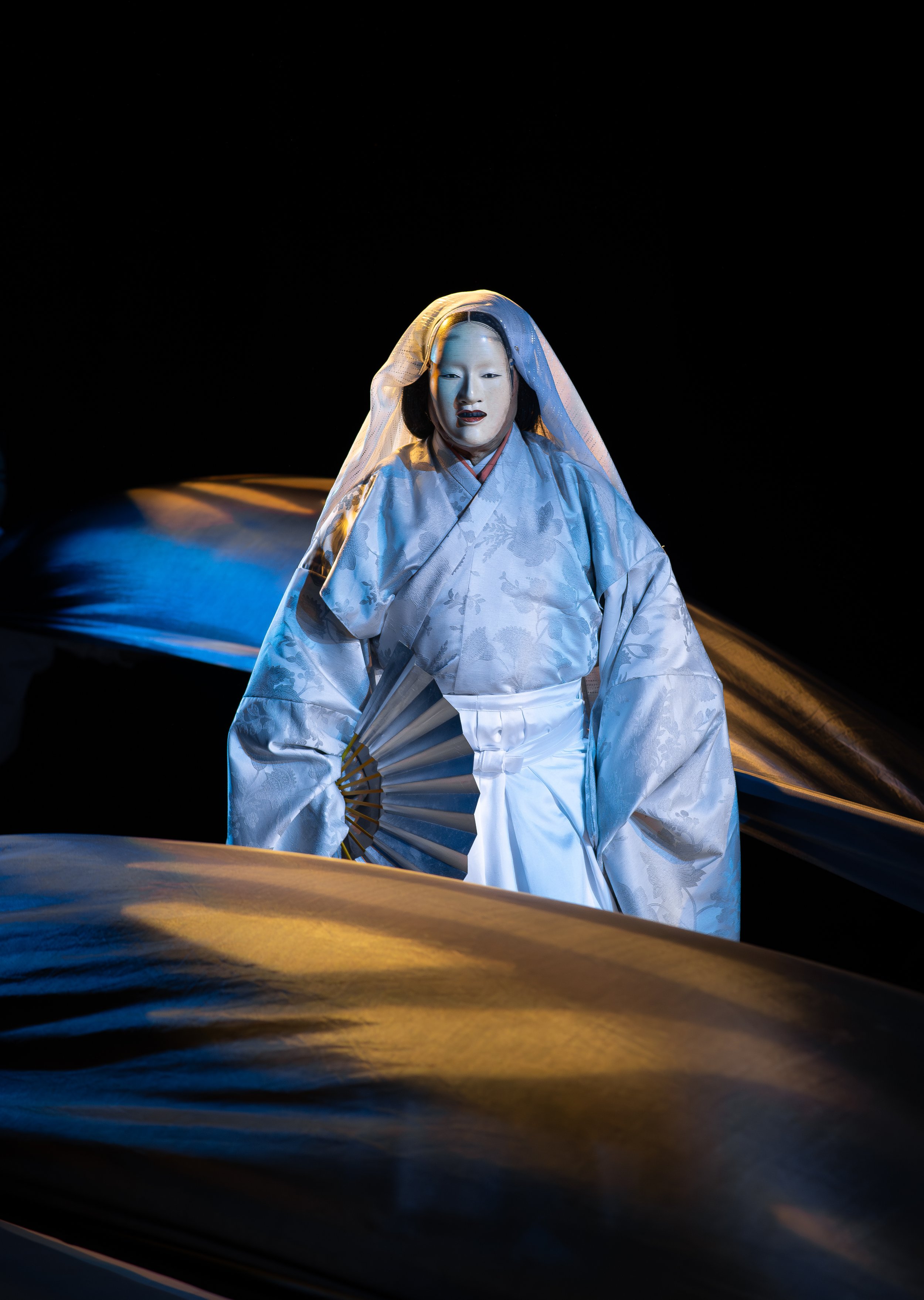 A performer in traditional Japanese costume with a white kimono, holding a folding fan, standing against a black background with flowing fabric.