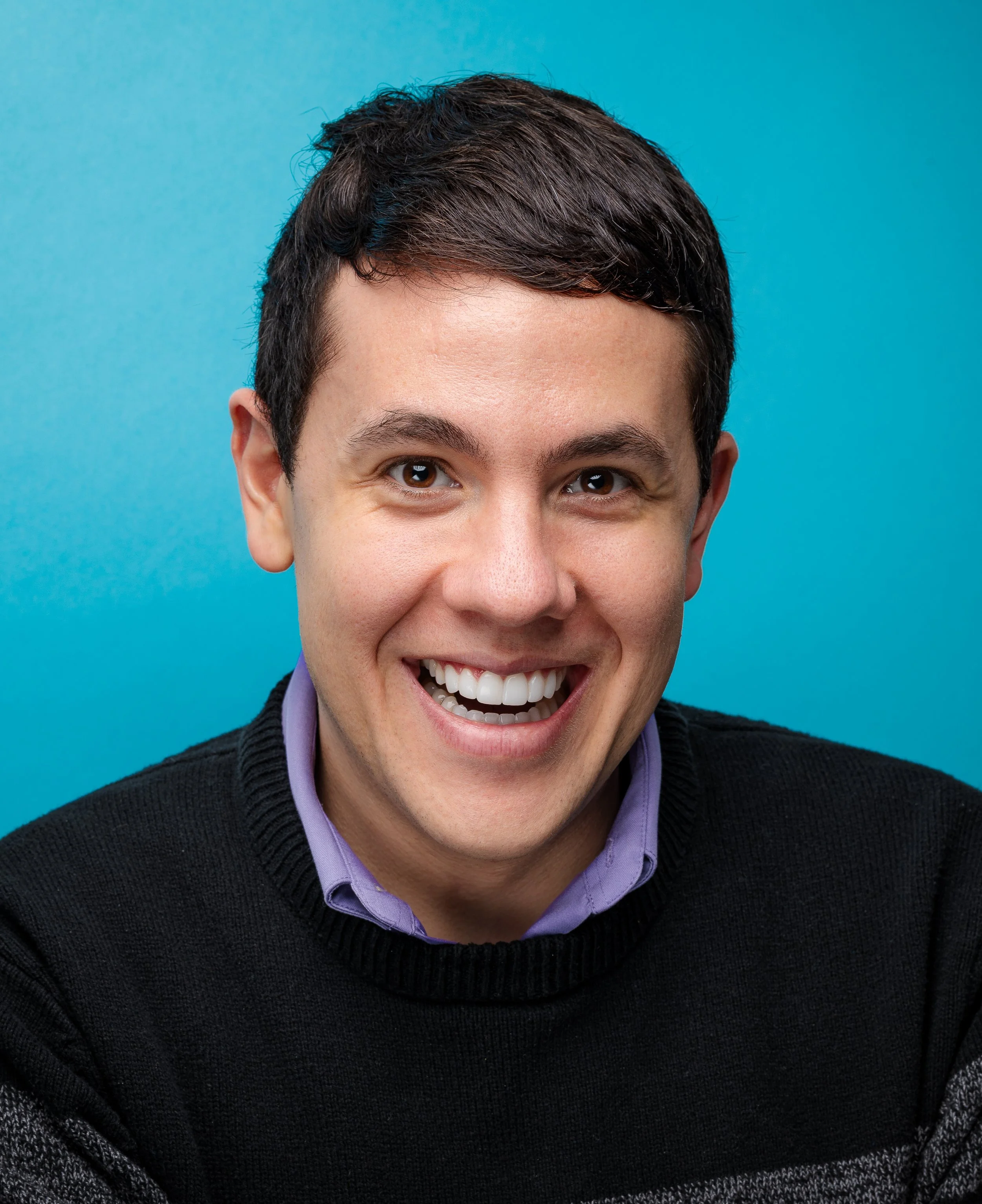 Close-up of a smiling man with dark hair, wearing a purple shirt and a black sweater, against a blue background.