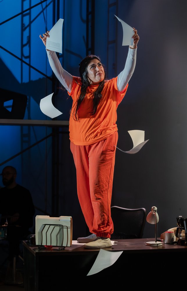 A woman standing on a desk with papers flying around in a theatrical setting.