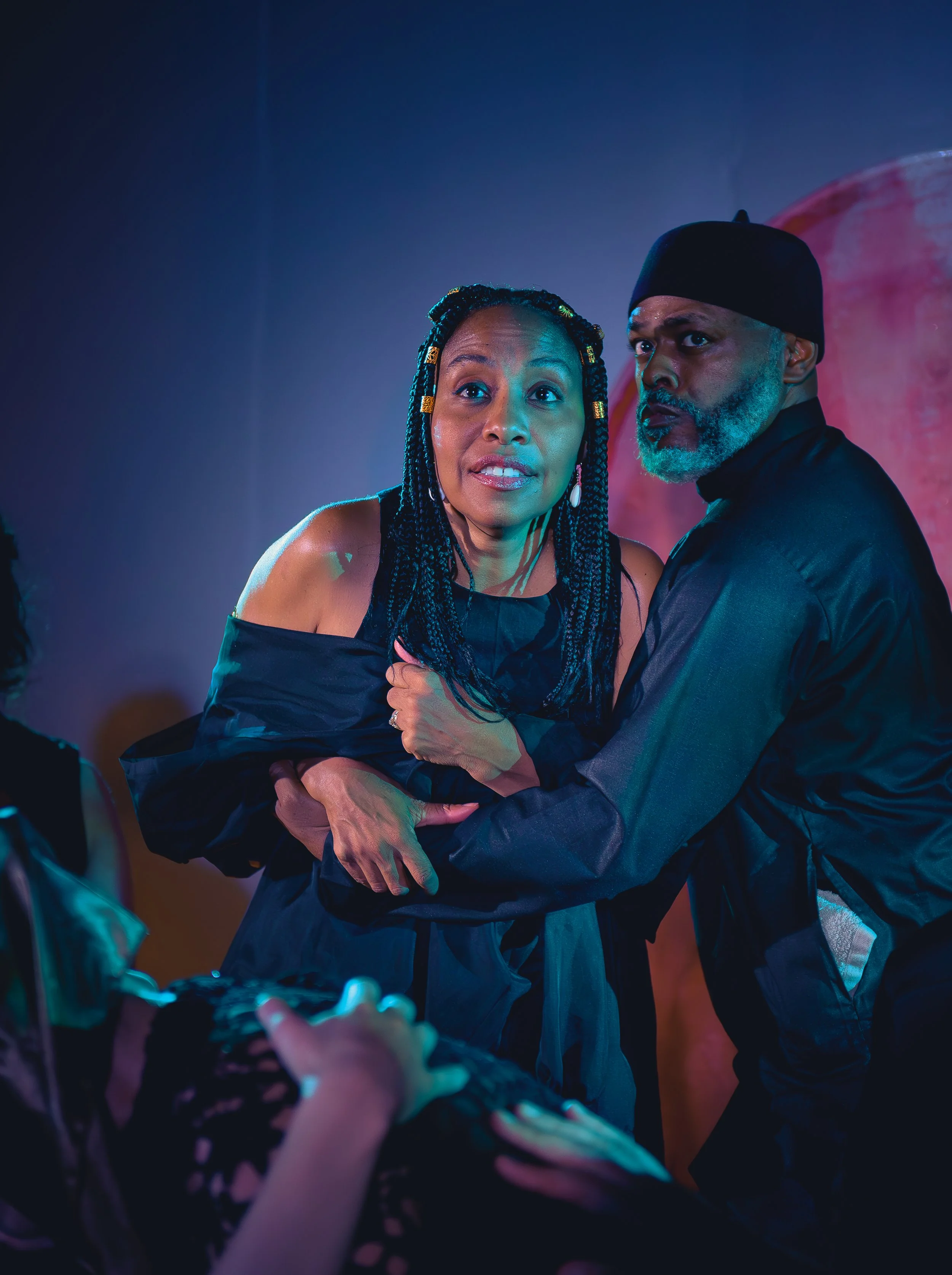 A woman with braided hair and a man with a beard and wearing a black hat are dancing together in a dark, colorful setting.