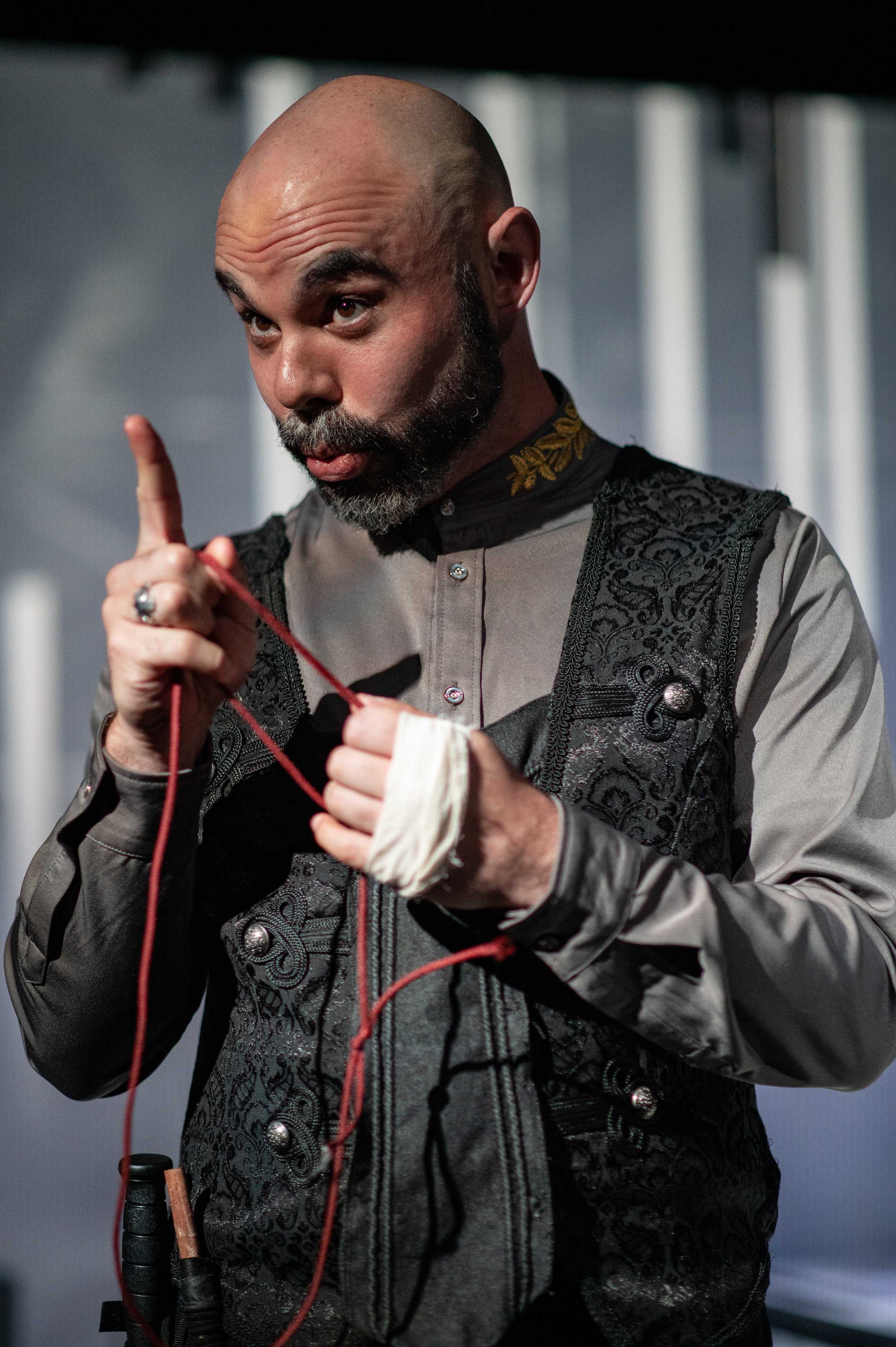 A man with a partially shaved head, beard, and dressed in a detailed gray and black embroidered vest is holding a winding bobbin with red thread. He is making a gesture with his index finger pointed upward, and appears focused on his task.