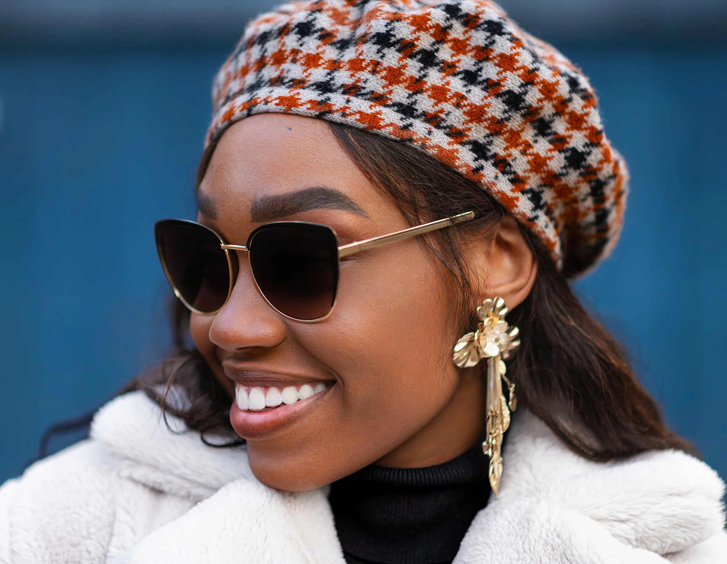Close-up of a woman smiling, wearing sunglasses, a patterned hat, and a white fuzzy coat over a black turtleneck against a blurred outdoor background.