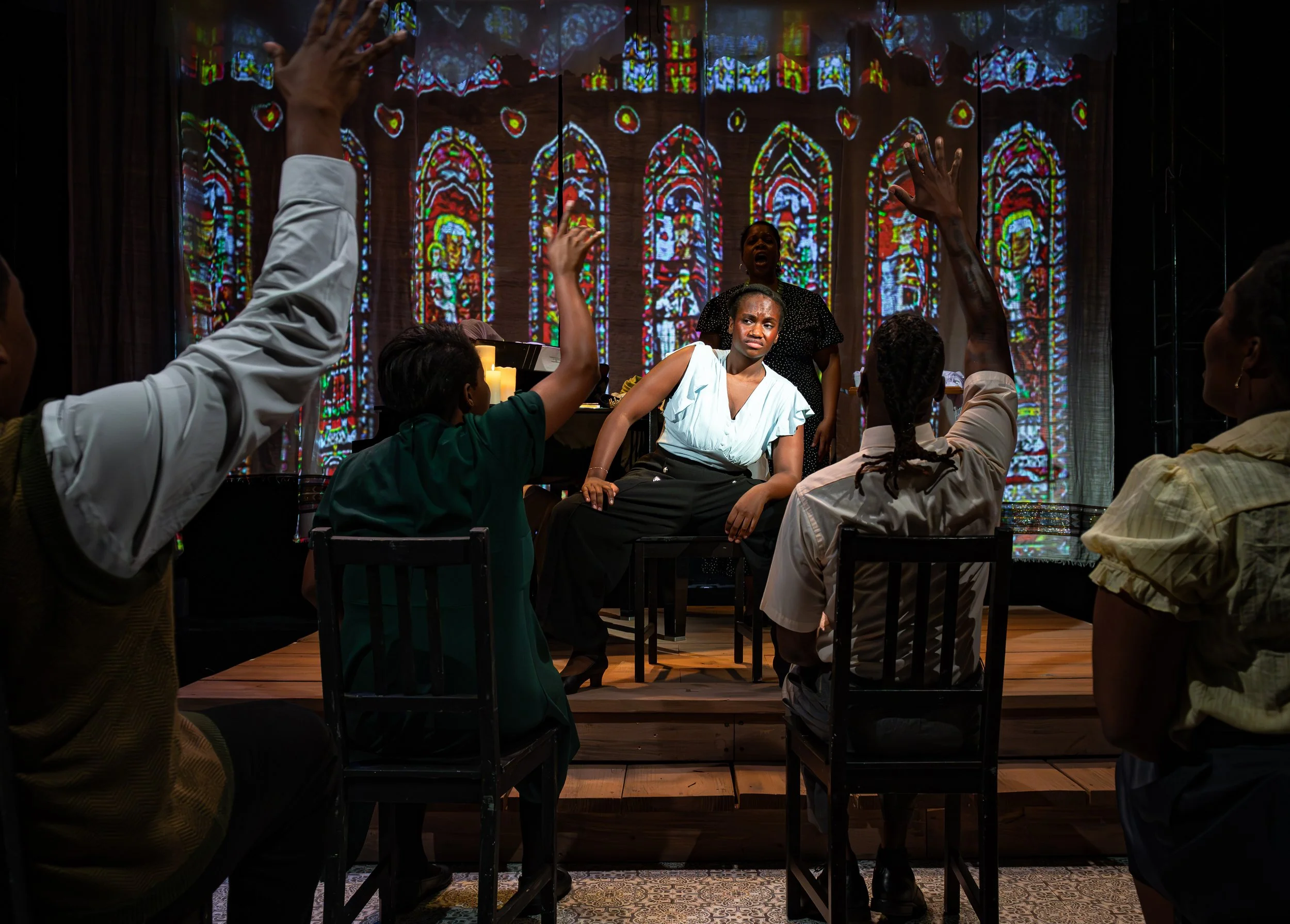 Group of people sitting at a table in front of stained glass window art, with three individuals raising hands, one woman with curly hair and a white top in the foreground, and candles on the table.