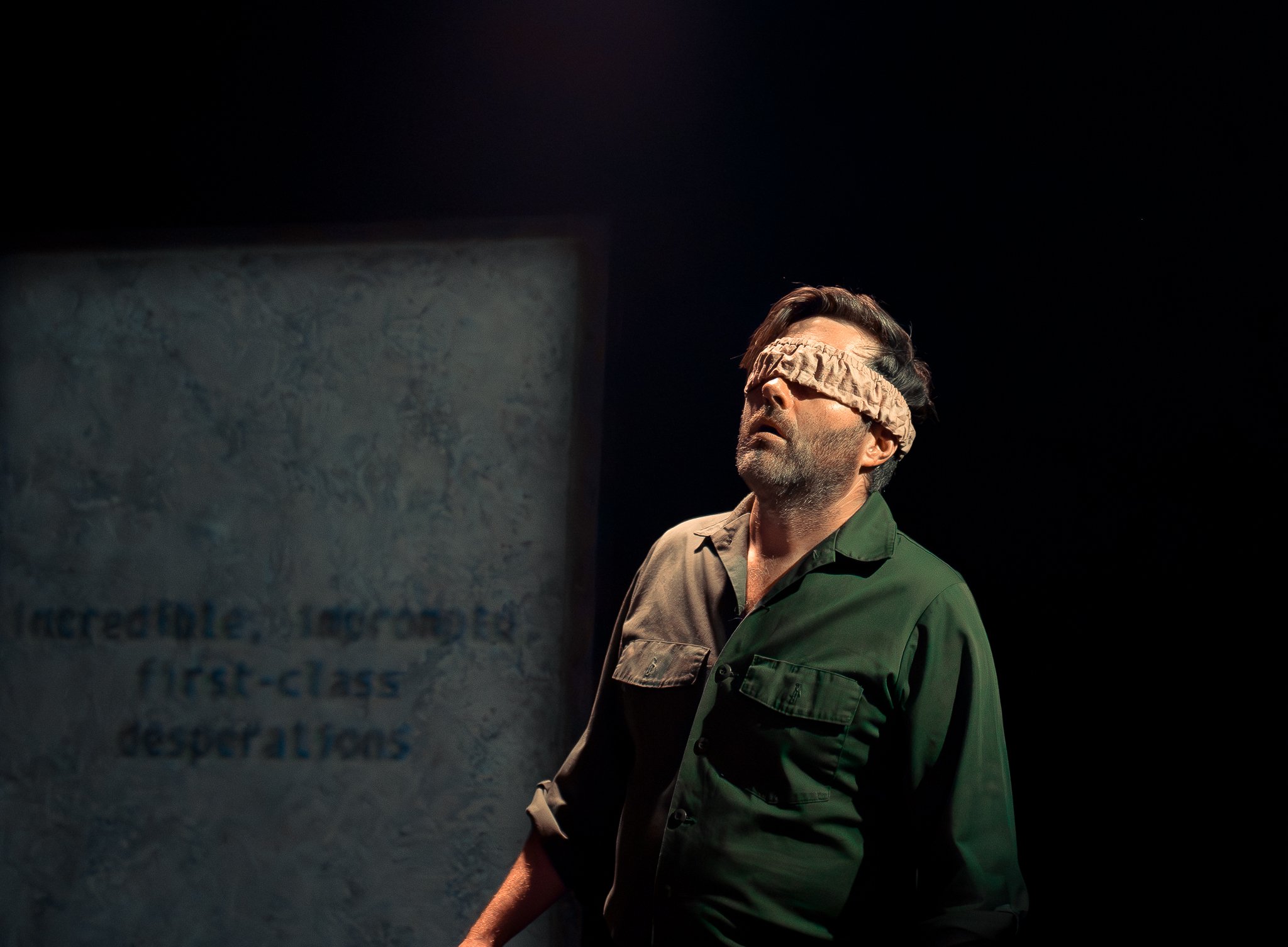 A man with dark hair and a beard wearing a green shirt and a blindfold.