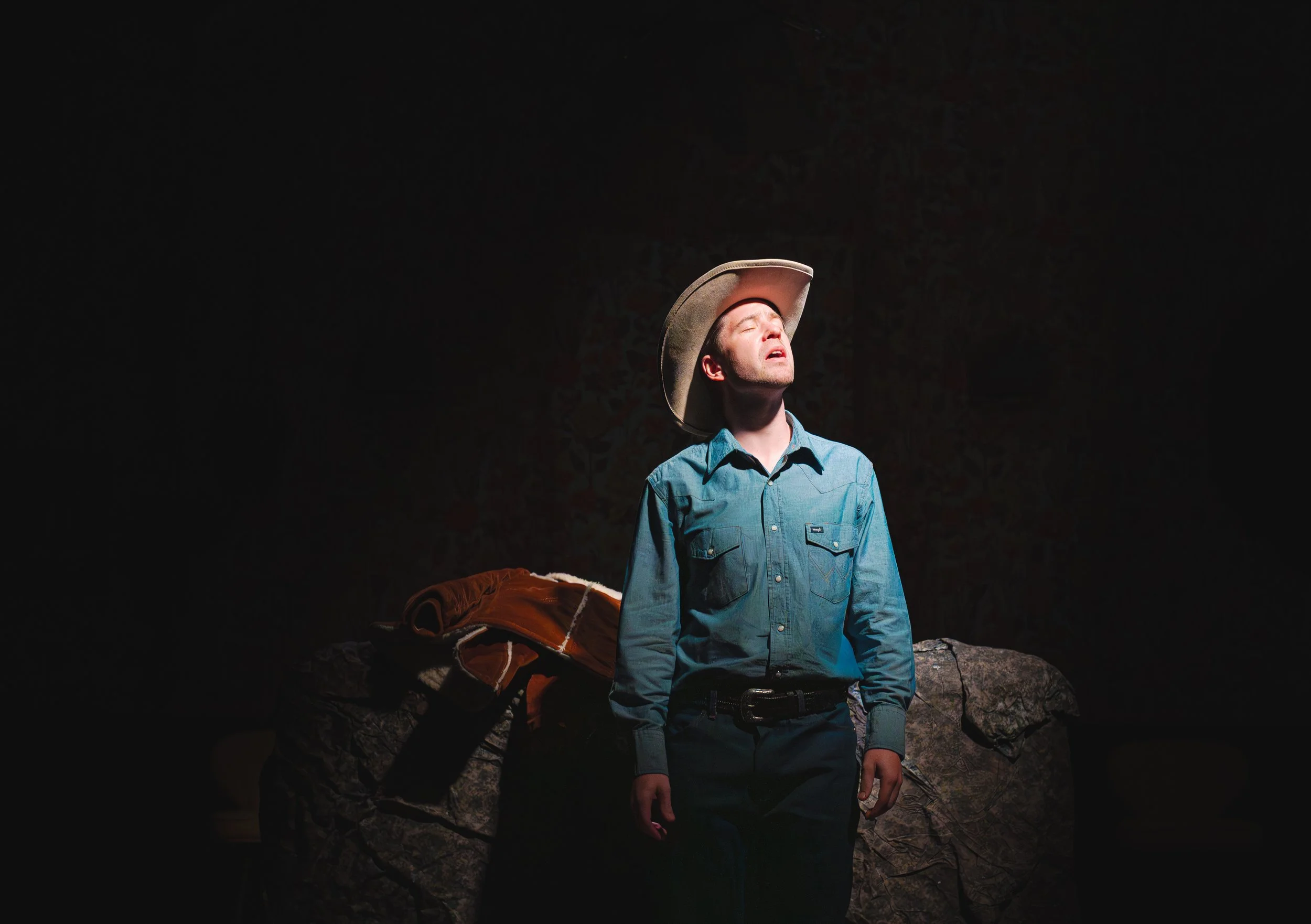 A man in a blue button-up shirt and cowboy hat stands on a rocky surface with a dark background. His eyes are closed and he appears to be enjoying the moment, with a blanket or jacket draped over a rock nearby.