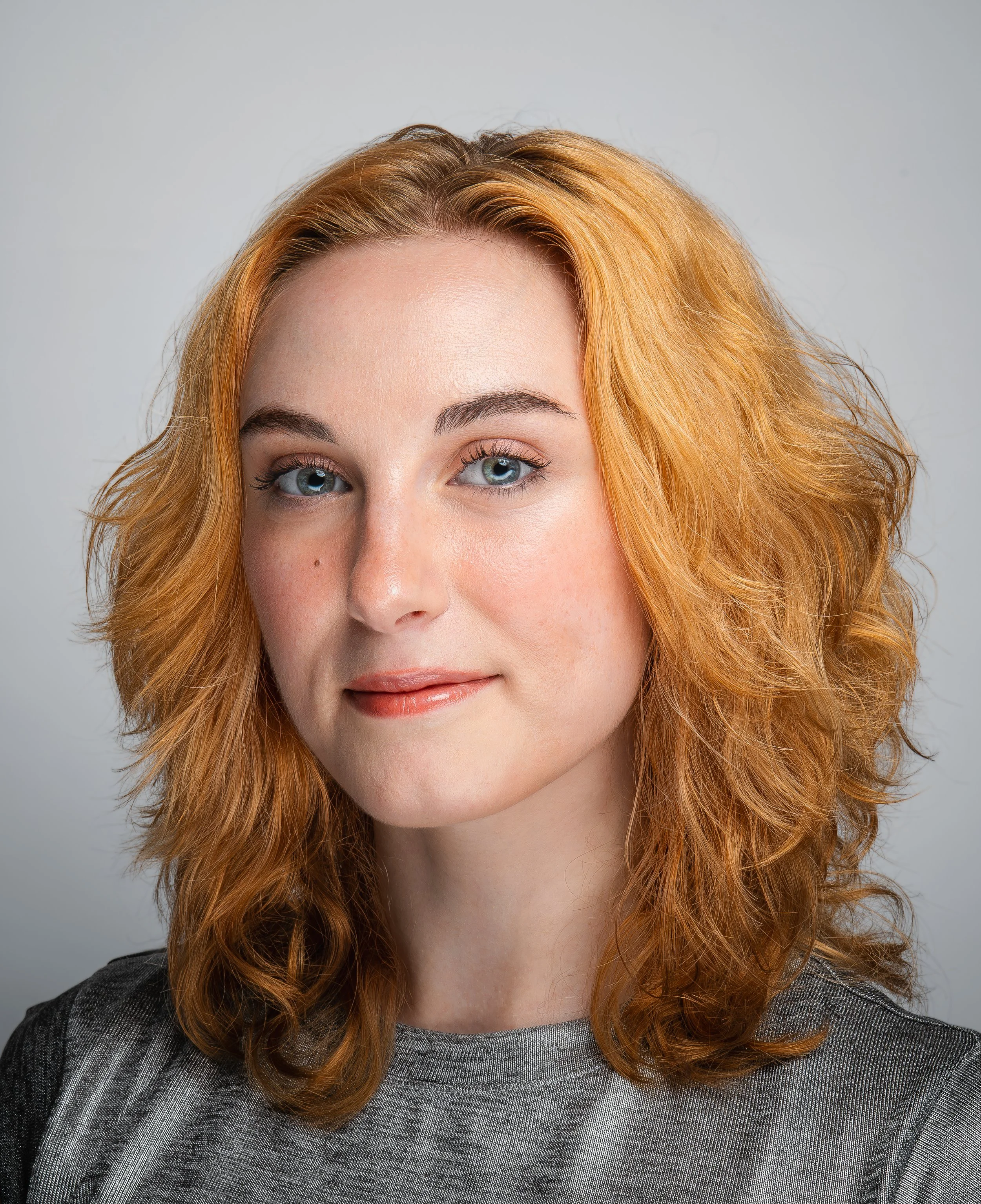 Close-up of a woman with curly red hair, blue eyes, and light makeup, wearing a dark gray textured top and standing against a plain light gray background.