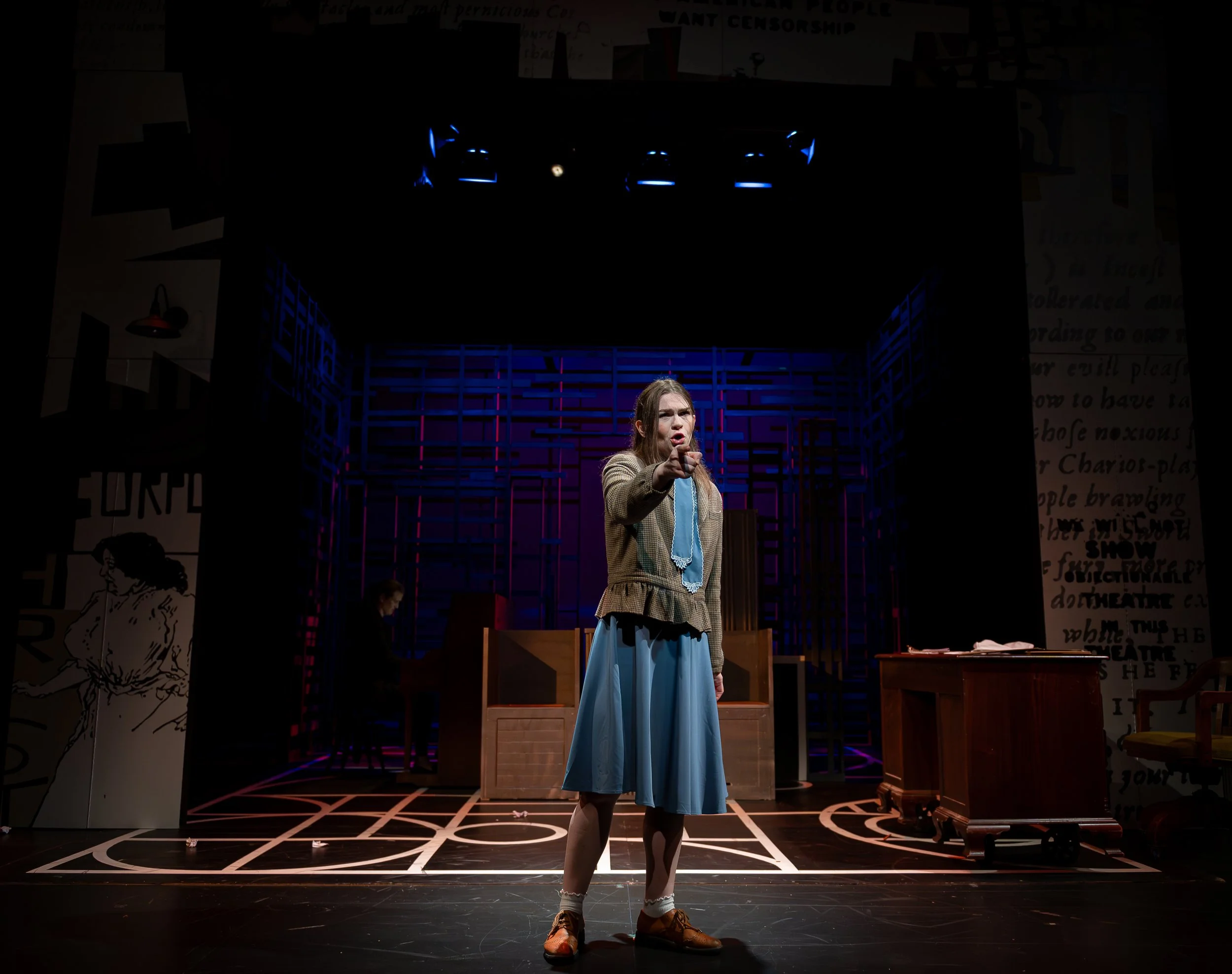 A woman performing on stage with a blue and beige costume, standing in front of a set with wooden furniture and colorful geometric background, under stage lighting.