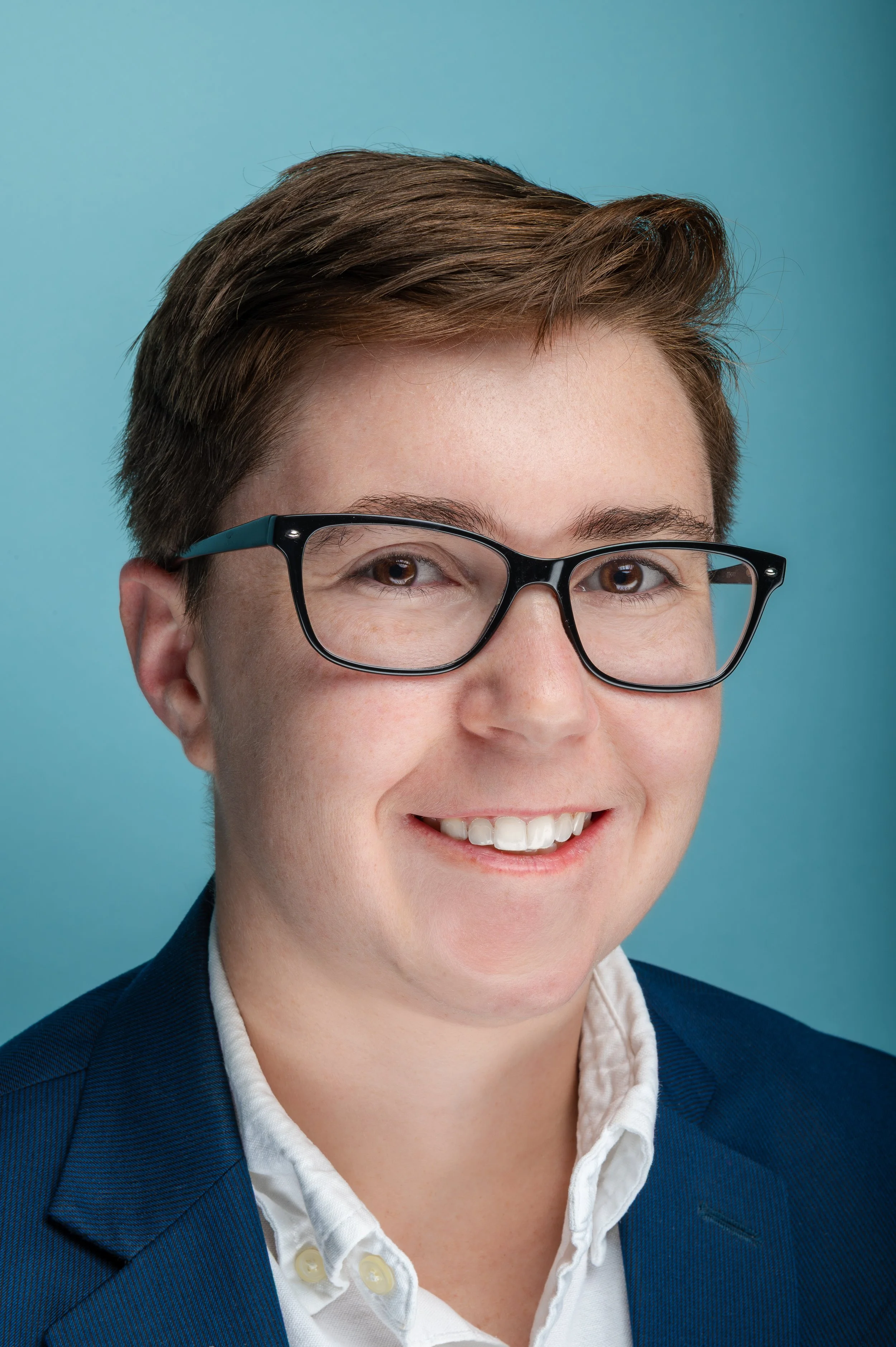 Close-up of a woman with short brown hair wearing black-rimmed glasses, a white shirt, and a dark blue blazer, smiling against a light blue background.