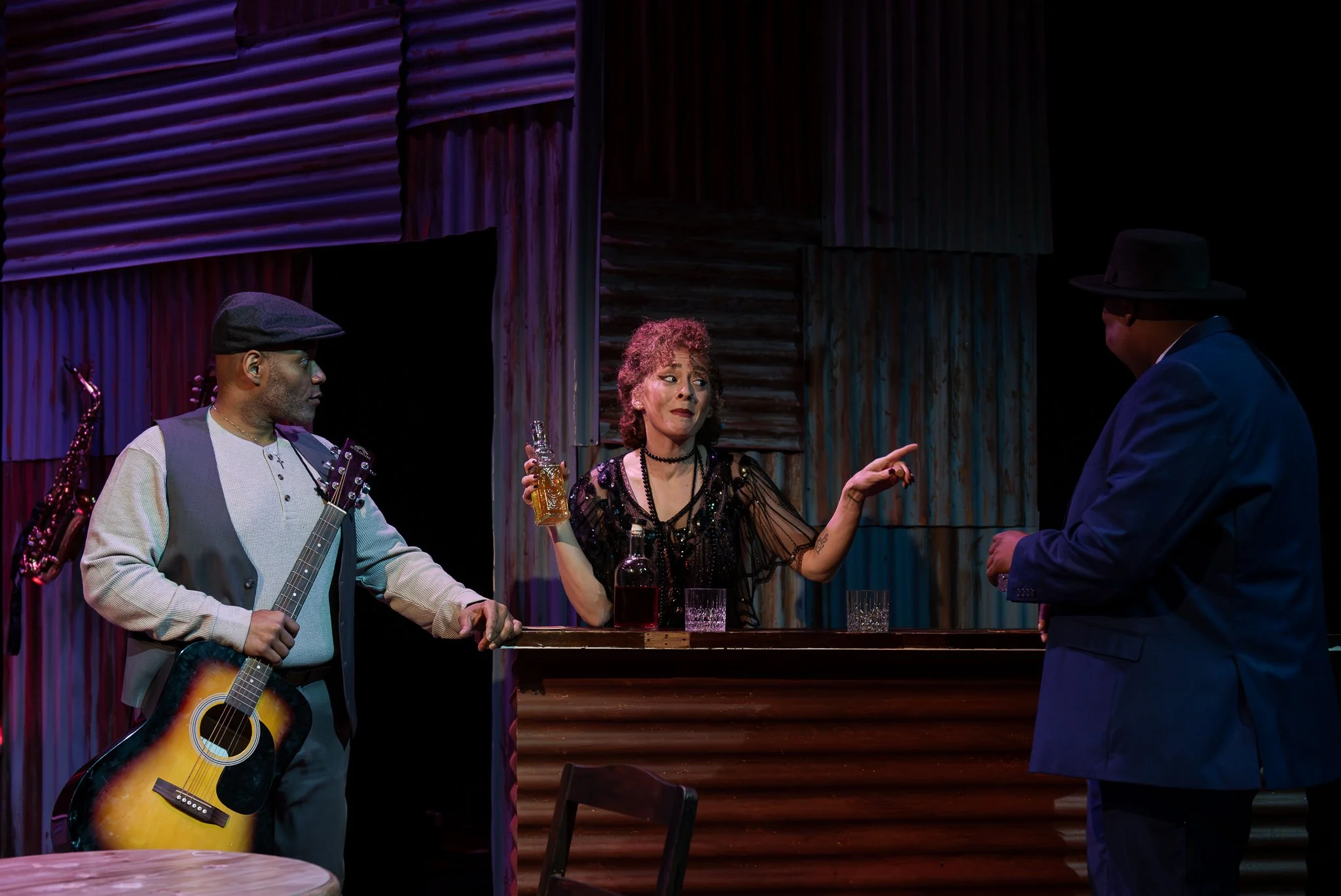 A scene from a stage play showing a woman with curly hair and a black dress, holding a bottle and gesturing while talking to two men. One man is playing an acoustic guitar and wearing a vest and cap, while the other man is wearing a dark suit and hat