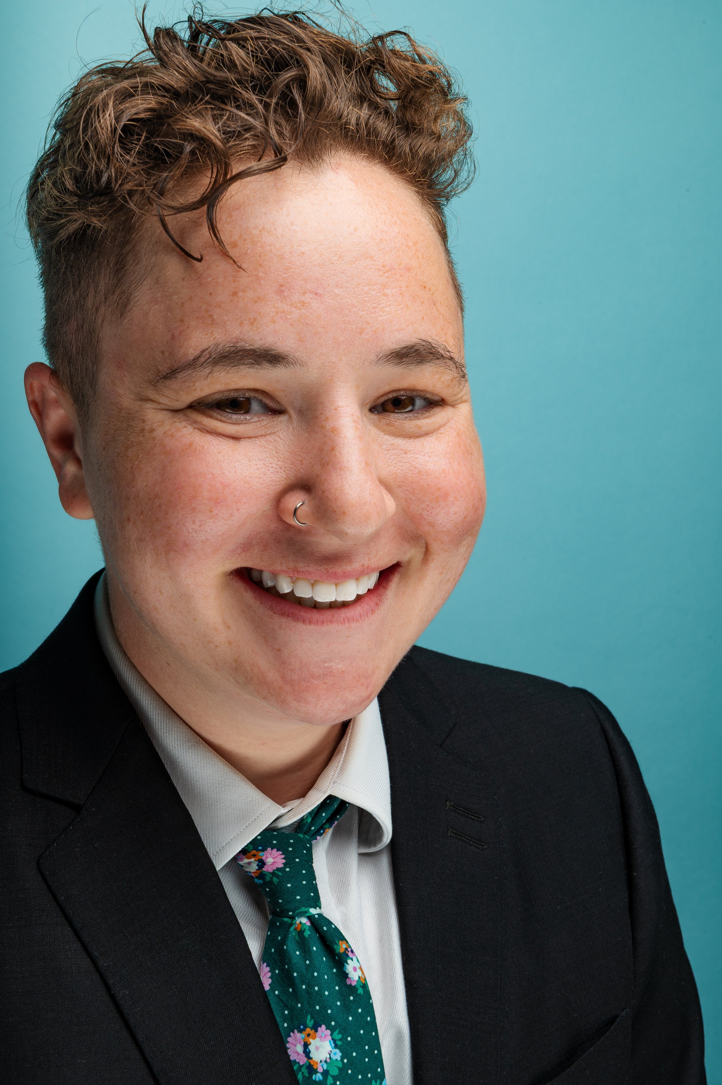 Close-up portrait of a person with short curly brown hair, wearing a black suit, white shirt, and featuring a nose ring, smiling against a light blue background.