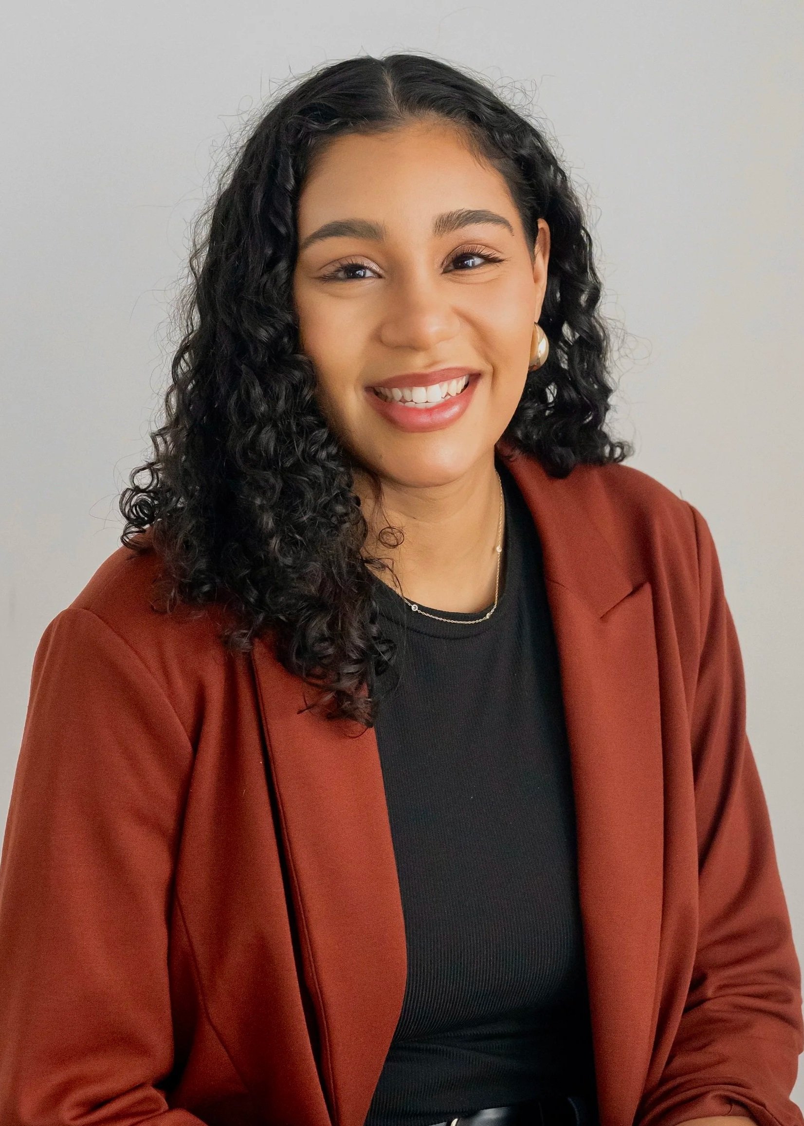 Woman with curly hair smiling outdoors, wearing a light blouse and a necklace with a 'C' pendant.