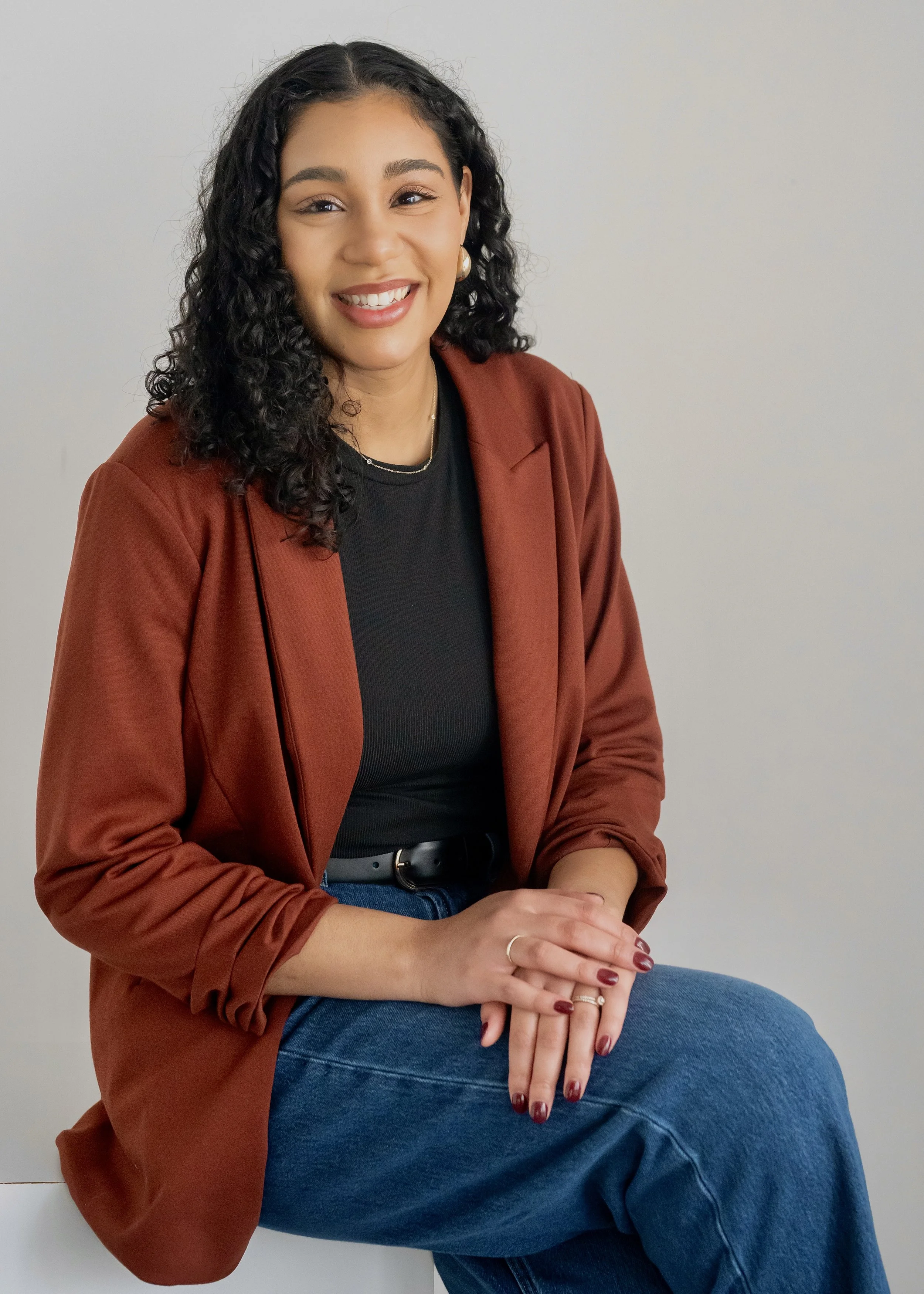 Woman with curly hair smiling, wearing a light-colored blouse and a "C" necklace, standing outside a building with large windows.