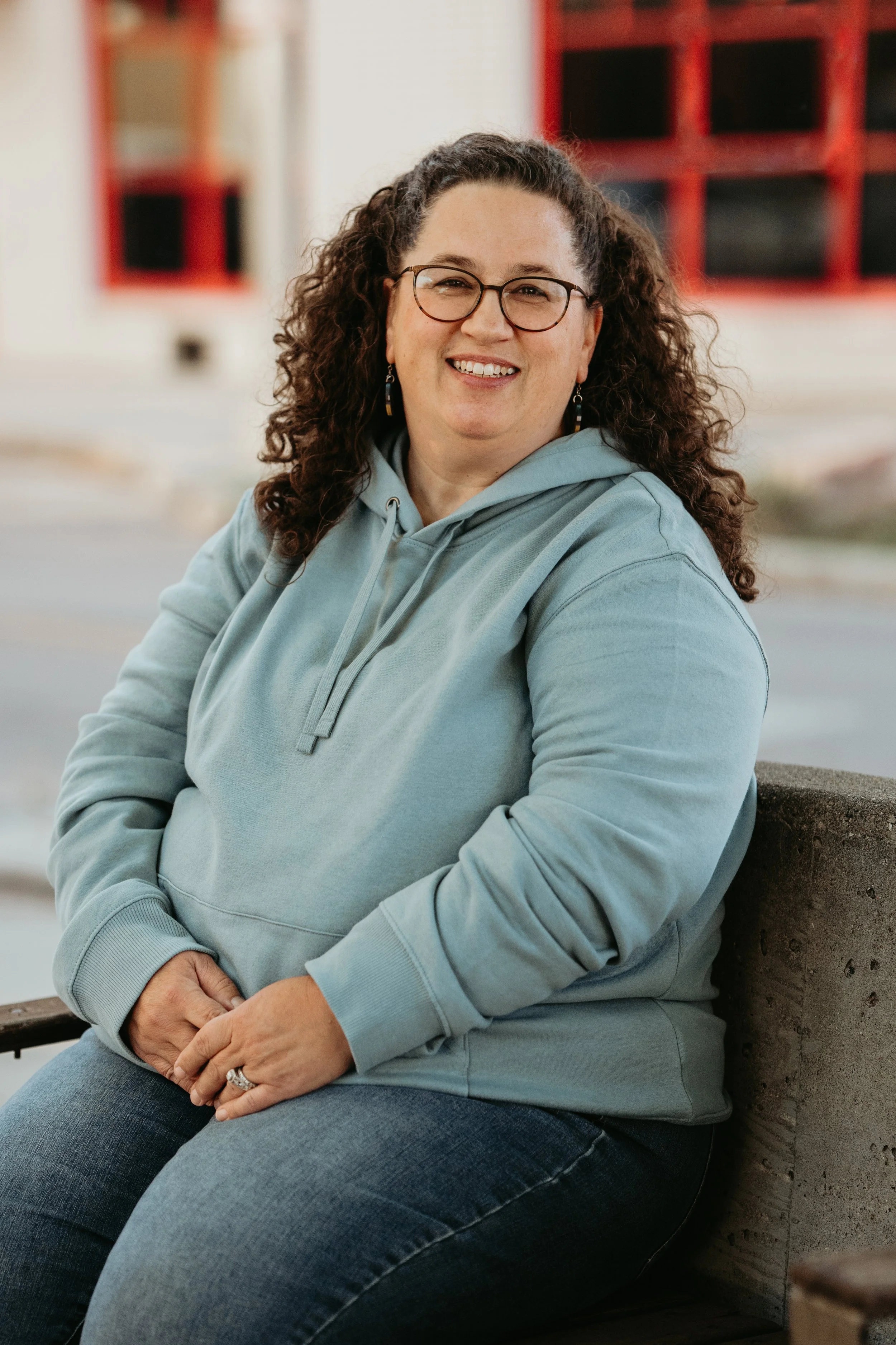 Smiling woman with curly hair wearing glasses and a blue hoodie seated outdoors.