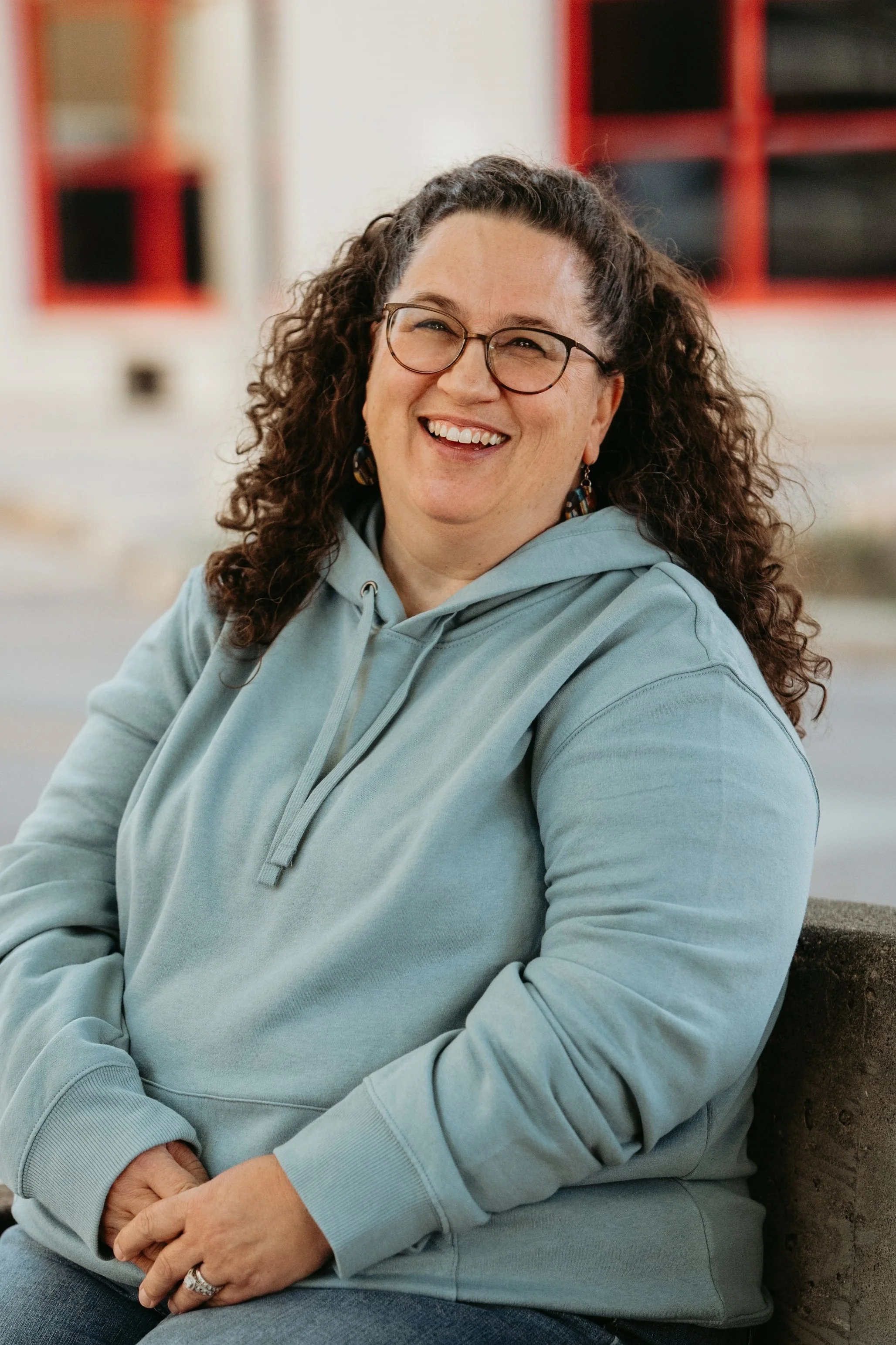 Smiling person with curly hair wearing glasses and a blue hoodie sitting outdoors.