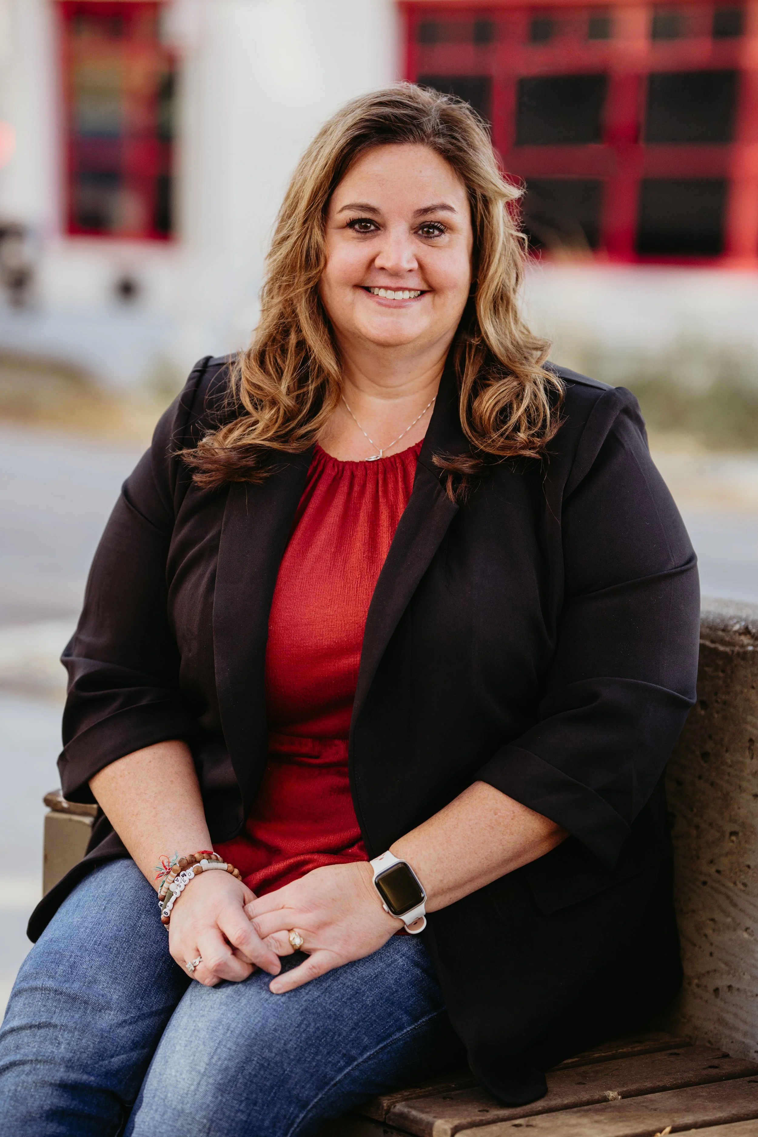 Smiling woman with wavy hair in a black blazer, red shirt, and jeans, wearing a smartwatch and bracelets, sitting outdoors on a bench.