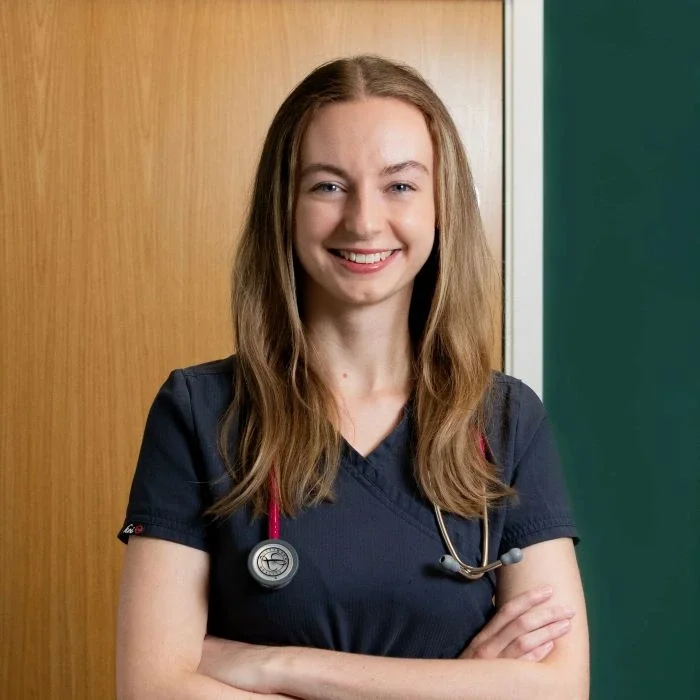 A smiling female healthcare professional standing indoors with arms crossed, wearing navy scrubs and a stethoscope.
