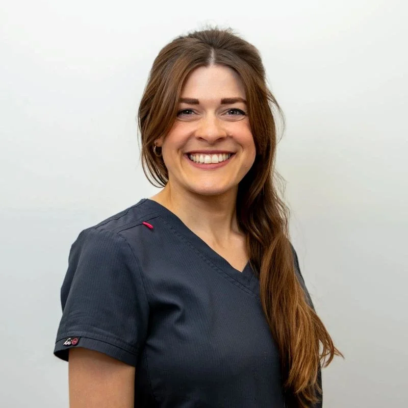 A smiling woman with long brown hair wearing navy medical scrubs standing against a plain white background.